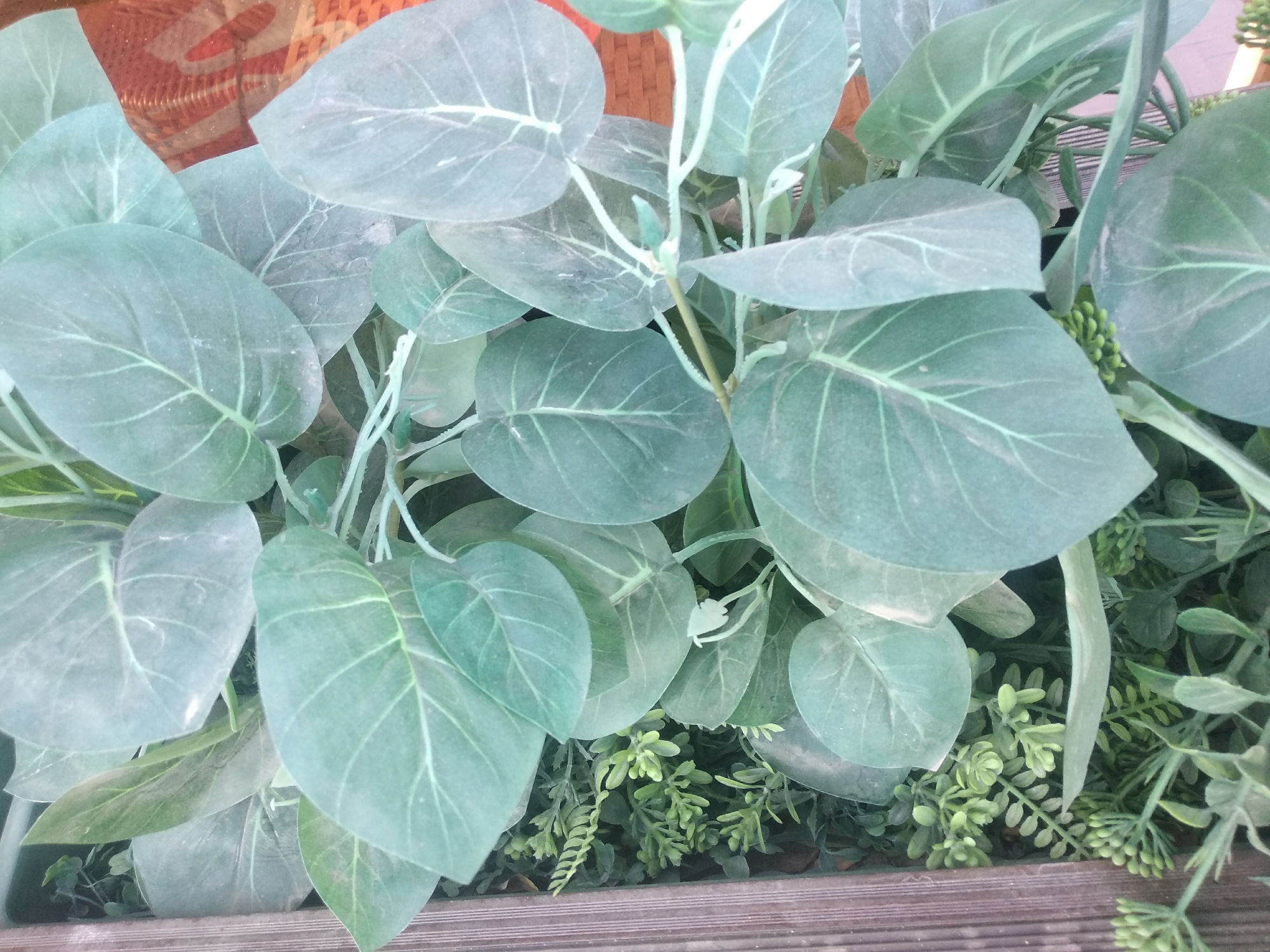 The image shows a close-up view of artificial eucalyptus leaves and other artificial greenery in a planter box. The planter box appears to be made of wood with horizontal slats. In the background, a woven textured surface, possibly a rug or textile, is visible. The overall color palette is dominated by shades of green and muted brown. The scene is an indoor or shaded outdoor setting, likely a home or commercial space, and does not provide any specific indicators to place it in Tashkent, Uzbekistan. There are no people or actions depicted.