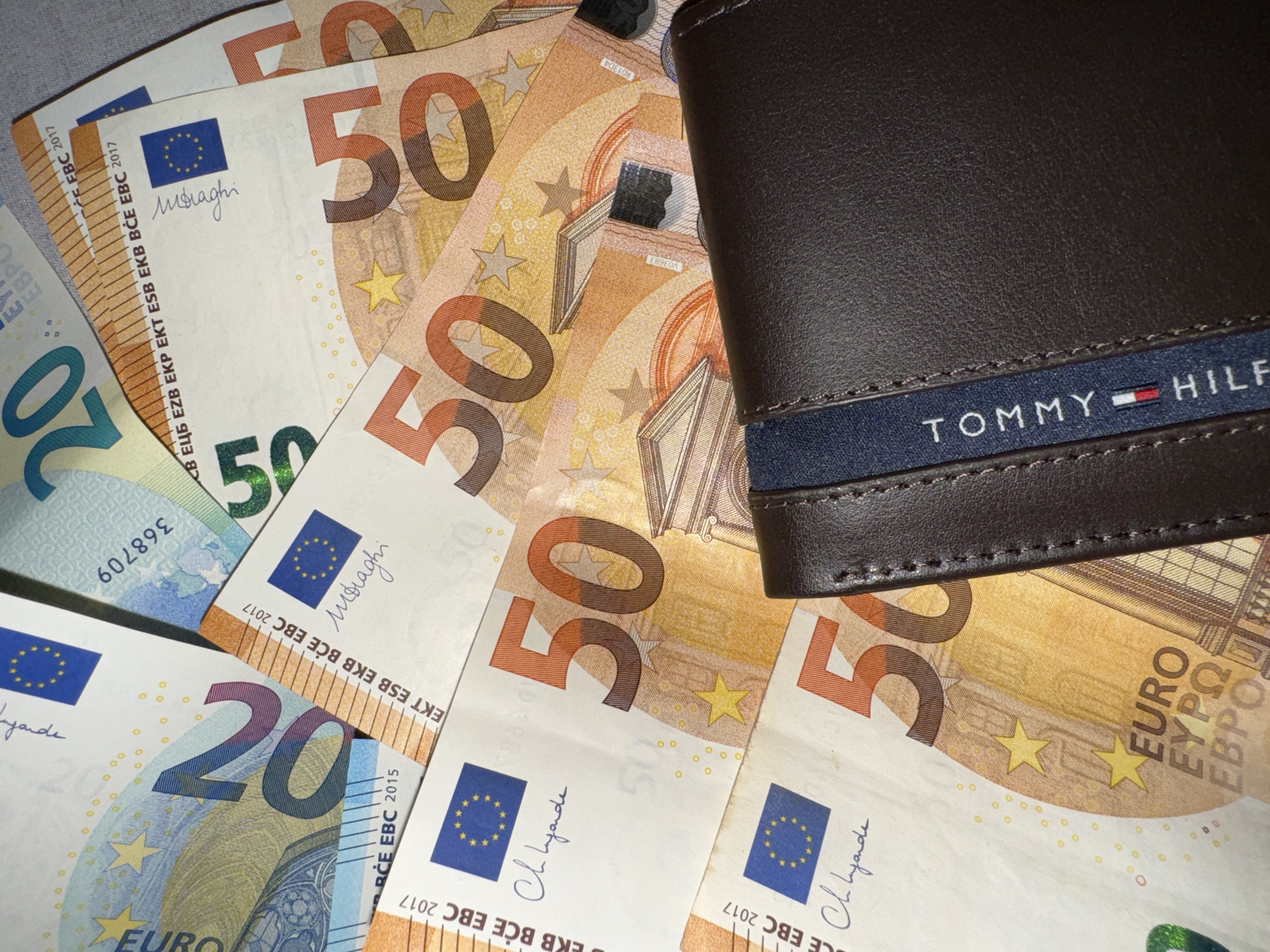 The image depicts a collection of Euro banknotes scattered around a brown Tommy Hilfiger wallet. There are several 50 Euro notes, recognizable by their orange color and the large number "50." Additionally, there are some 20 Euro notes present, which have a blue color scheme and the number "20." The banknotes are partly obscured by the wallet. The scene takes place in an indoor location, possibly an office or living room.