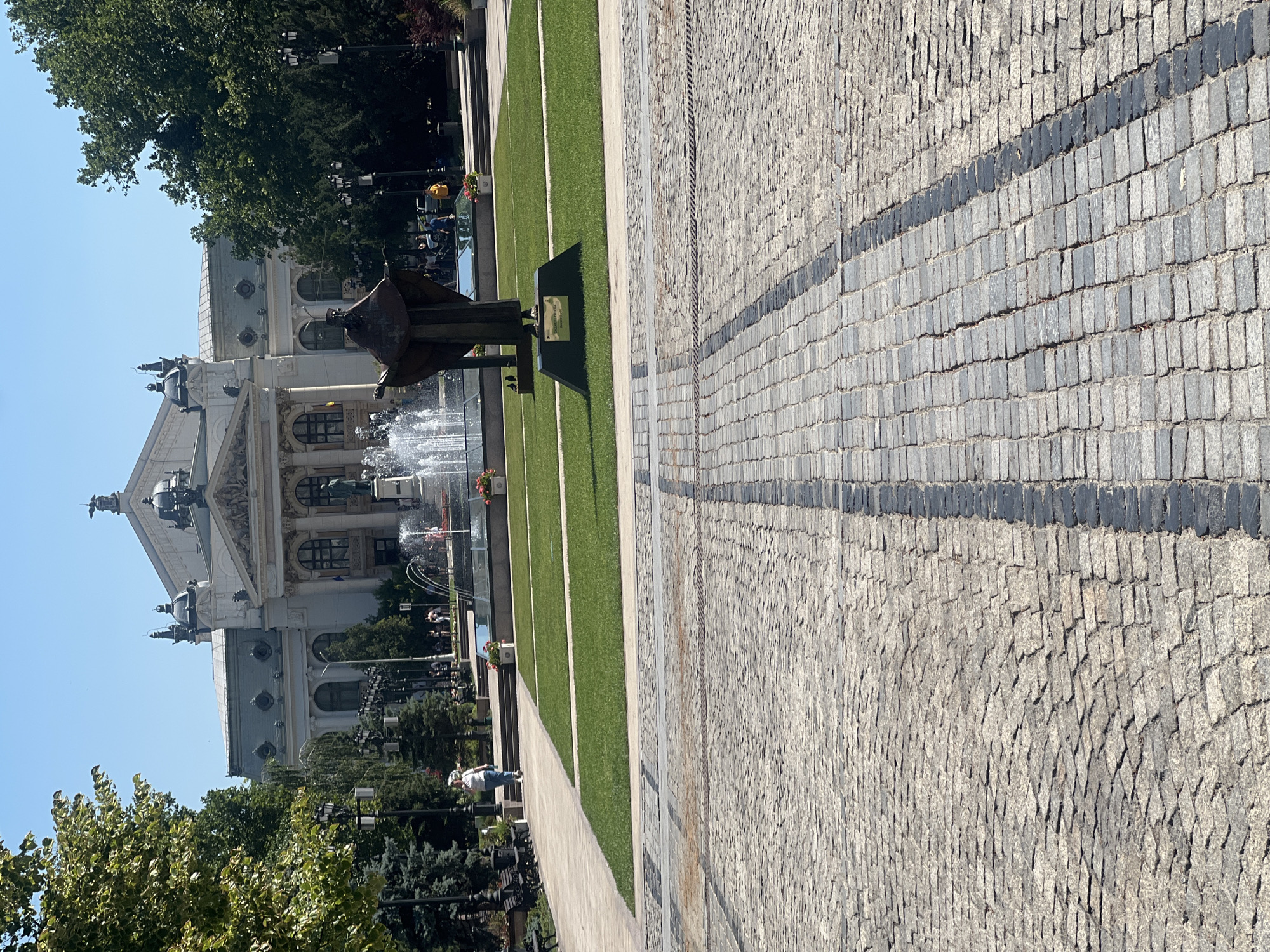 The image shows a wide, paved walkway with a distinct pattern of cobblestones and dark grey stone strips. To the left of the walkway is a manicured lawn area with a fountain and a bronze statue. Behind the lawn is a grand, ornate building with classical architecture, featuring columns, intricate carvings, and domes. Trees with lush green foliage line the left side of the scene, and in the background, more trees and buildings are visible. Several people are scattered throughout the scene. Some are walking along the paved path, while others are gathered near the fountain or in front of the building. One person is visible on the lawn, and a few are seated in the background. The time of day appears to be daytime, with clear blue skies indicating fair weather. The scene evokes a sense of a public space, possibly a park or a plaza in front of a significant cultural or governmental building. The overall atmosphere is calm and leisurely, suggesting a typical day in a city. No visible text is present in the image.