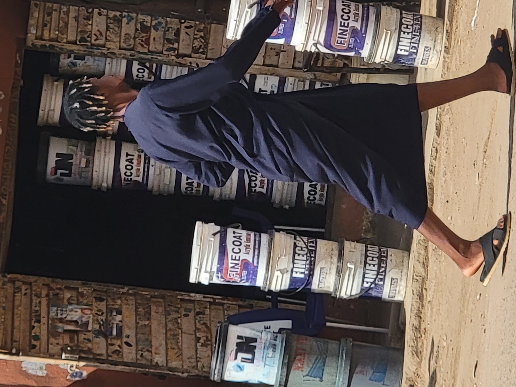 A person with dreadlocks is standing in front of a small shop stacked with buckets of "FINECOAT" acrylic emulsion paint. The person is wearing a dark blue, long-sleeved garment and black sandals. The shop appears to be in a somewhat worn condition, as indicated by the wooden beams and visible posters on the wall. The setting is outdoors, with a sandy ground or sidewalk visible. The location is Maiburiji, Nigeria.