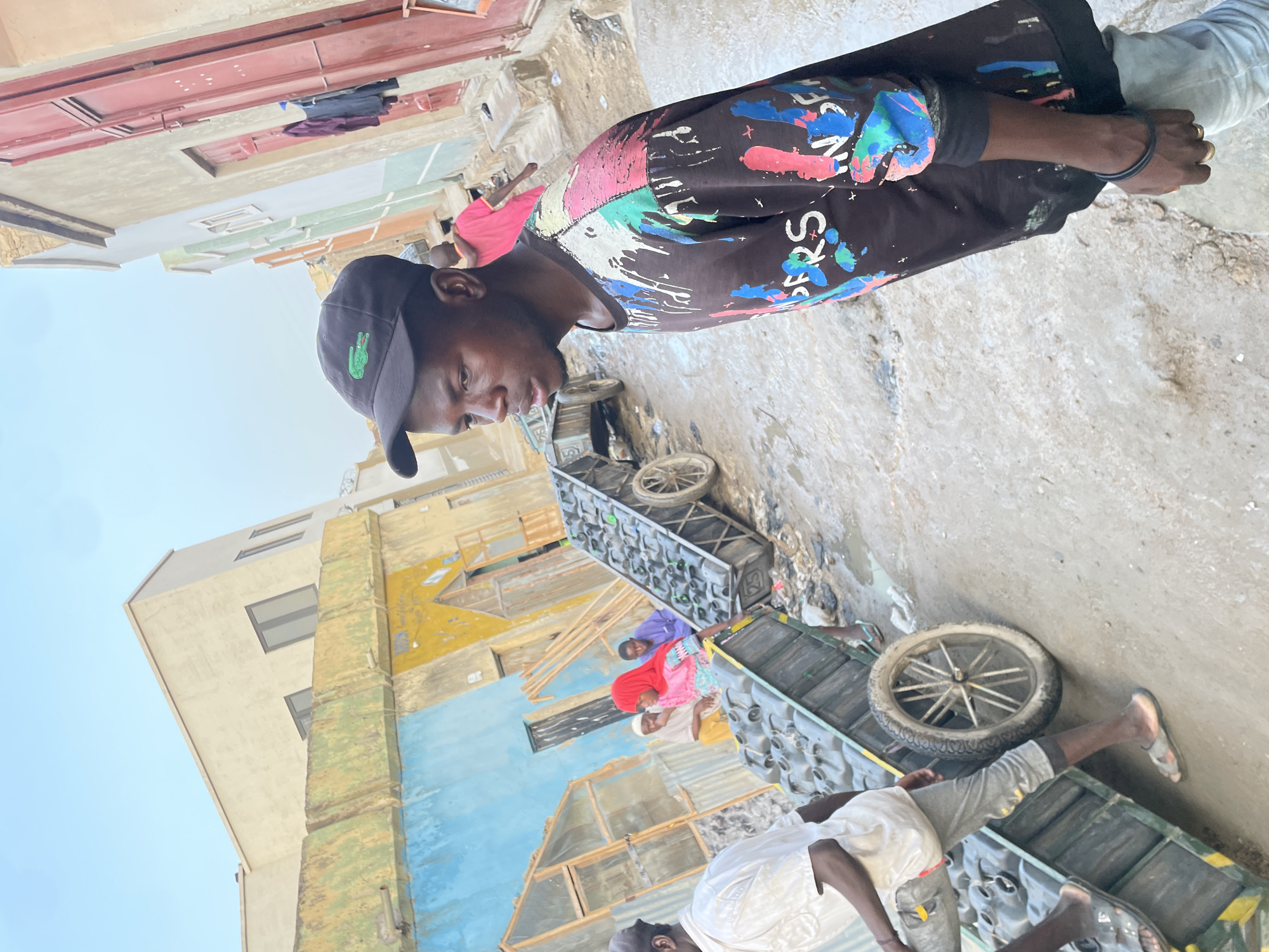 A man wearing a black shirt with colorful designs and a black baseball cap is looking down at a group of people transporting items on wheeled carts on a street in an urban environment. The street is flanked by buildings with colorful facades, including blue, yellow, and beige tones. The wheeled carts appear to be carrying what might be bottles or similar items. The environment suggests an impoverished area within the city of Unknown, Unknown.