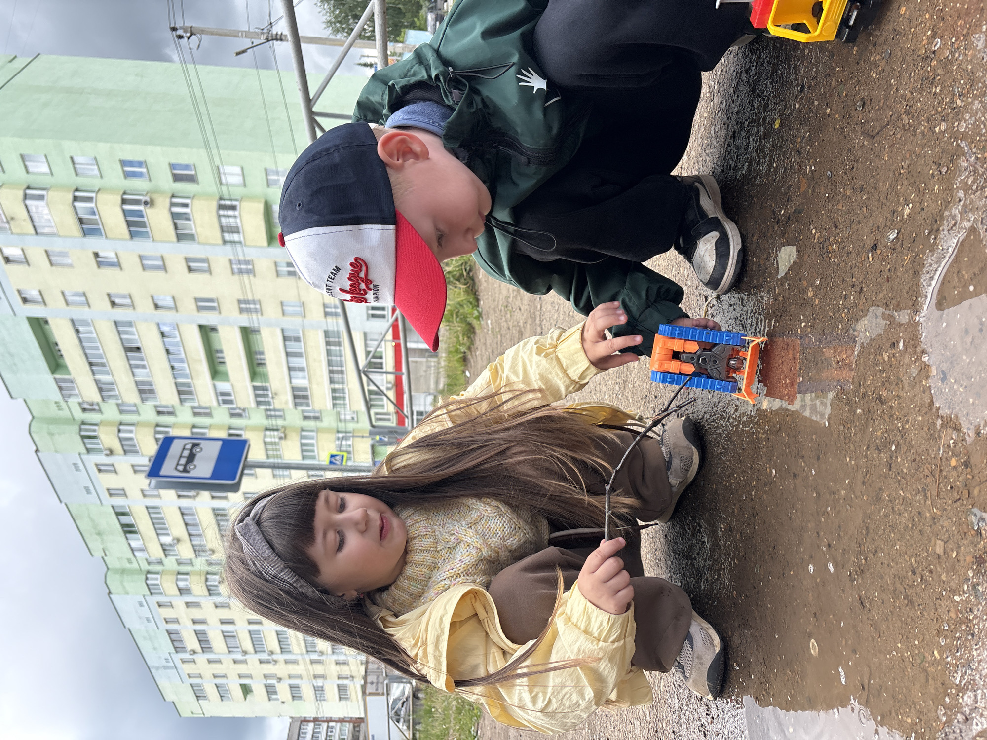 Two children are engrossed in play on a wet, gravelly asphalt surface next to a large puddle, likely after recent rain. The image, set in an urban residential area of Chaikovsky, Russia, shows multi-story buildings with light green and yellow facades under an overcast sky in the background.
On the left, a boy in a dark green jacket, black pants, and sneakers crouches down. He wears a baseball cap that is dark blue, white, and red, with visible text reading "ELENT TEAM," "I league," and "CHAMPION." He is looking intently at a blue and orange toy vehicle positioned in the puddle.
On the right, a girl with long dark hair and a plaid headband is also squatting. She wears a light yellow long-sleeved shirt over a colorful knitted sweater and brown pants, with light sneakers. She holds a thin stick with both hands, also focused on the puddle area. Another yellow and red toy truck is partially visible near the boy's feet, at the edge of the puddle.
Behind the children, a blue bus stop sign featuring a white bus icon is clearly visible. The overall scene captures a moment of focused outdoor play, with the children seemingly unfazed by the damp conditions, enjoying their toys and the puddle.