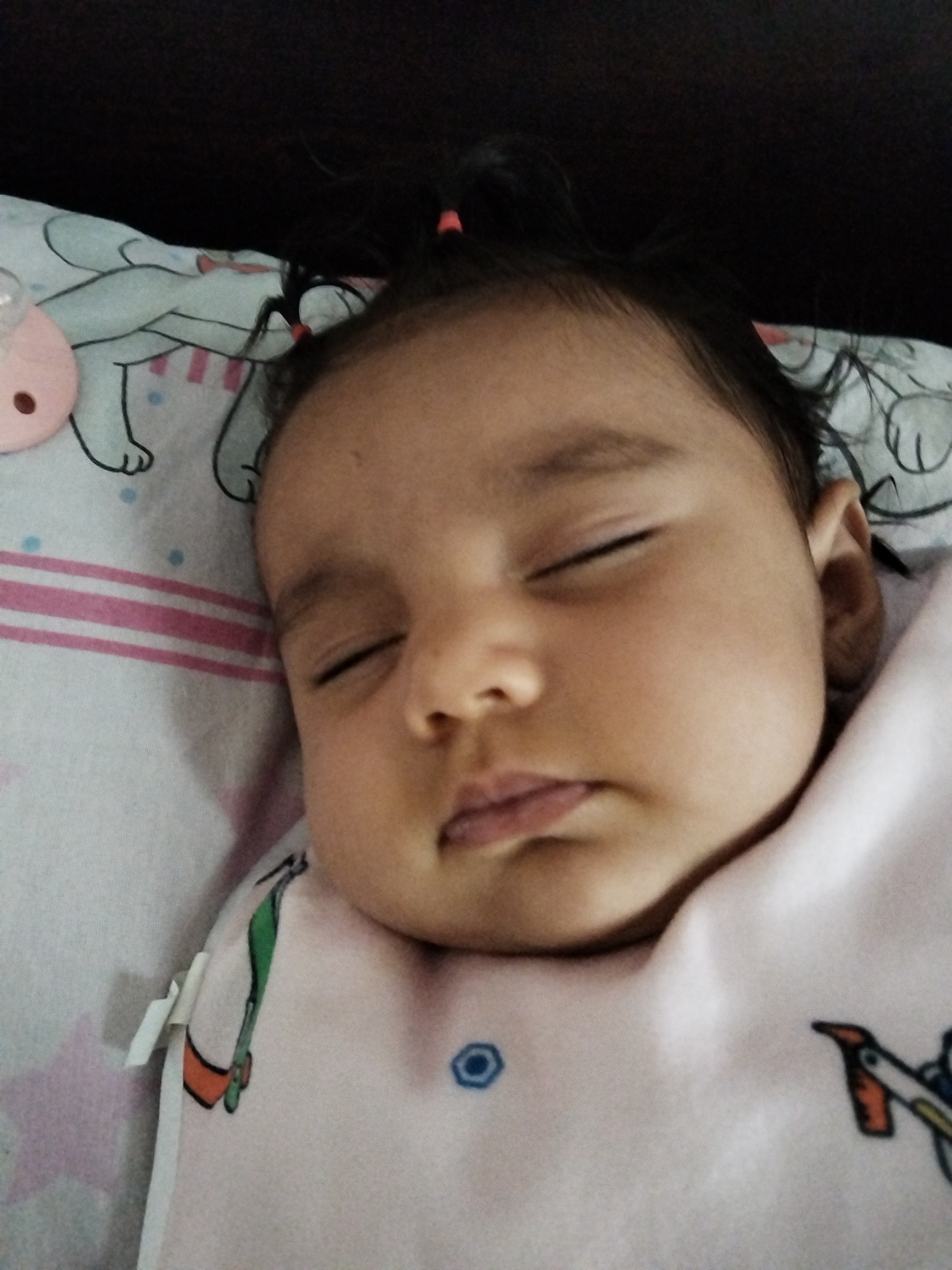 A close-up shot depicts a baby sleeping. The baby is lying on a pillow with a pink and white striped pattern and a cartoon character illustration. The baby is wearing a light pink garment with a blue button and a penguin-like design. A pacifier is visible near the pillow. The setting appears to be indoors.