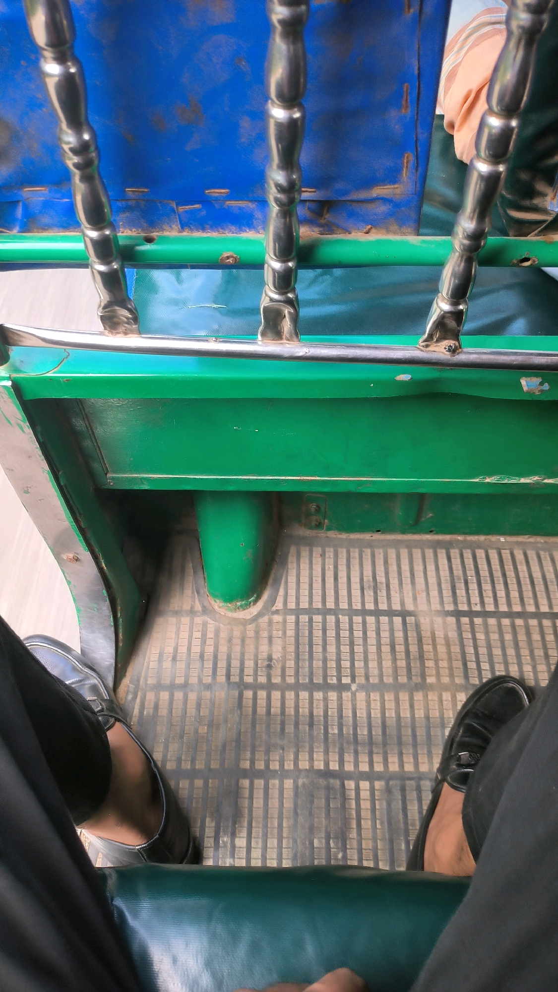 The image shows the interior of a vehicle, likely a rickshaw or a small auto-rickshaw, seen from the perspective of a passenger. The floor is covered with a ribbed, grey mat. Two pairs of legs are visible, one on the left wearing a black shoe and the other on the right wearing a black loafer. The passenger on the left has dark skin and is wearing dark trousers and a dark shoe. The passenger on the right also has dark skin and is wearing dark trousers and a black loafer.
The vehicle's interior features a prominent green metal seat or compartment. Above this, there are chrome-plated decorative posts and a bright blue tarp, possibly the roof or a side panel. A person's torso wearing an orange and white striped shirt is partially visible in the upper right corner, suggesting another passenger or the driver.
The lighting suggests it is daytime, and the scene is likely taking place on a street in Begumganj Upazila, Bangladesh. The overall impression is of a mode of transport commonly found in this region.