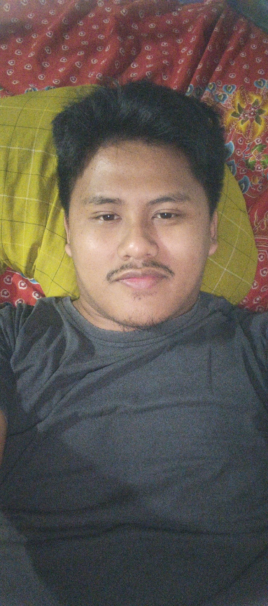 A close-up, head-and-shoulders selfie is shown, taken from a slightly elevated angle. The subject is a male with dark hair and a mustache, wearing a dark gray t-shirt. He is lying down on a bed with a yellow patterned pillow behind his head, and a red patterned blanket under the pillow. The image appears to be taken indoors in Kemuning, Indonesia.