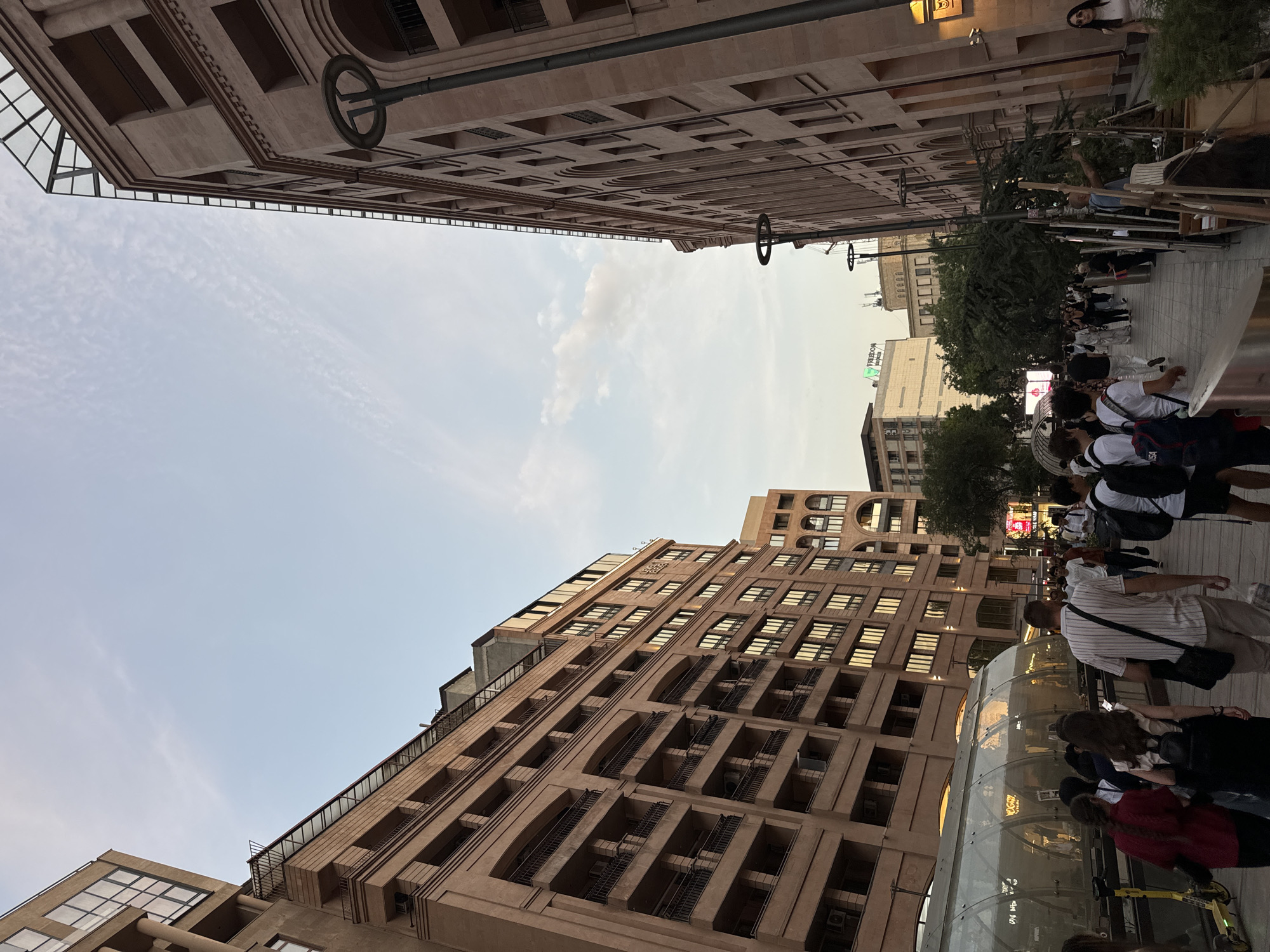 The image depicts a street scene in Yerevan, Armenia, likely during the late afternoon or early evening given the lighting. On the left, a large, multi-story building with a reddish-brown facade and numerous balconies dominates the view, with a glass-covered structure at street level that appears to be an entrance to an underground passage. To the right, another similar building with arched windows and a modern glass roof extension rises into the sky. A crowd of people is walking along the pedestrianized street, some carrying bags and backpacks. A yellow electric scooter is visible near the glass structure on the left. Trees and streetlights line the pathway. The sky above is a pale blue with wispy clouds. The overall impression is one of a bustling urban environment.
