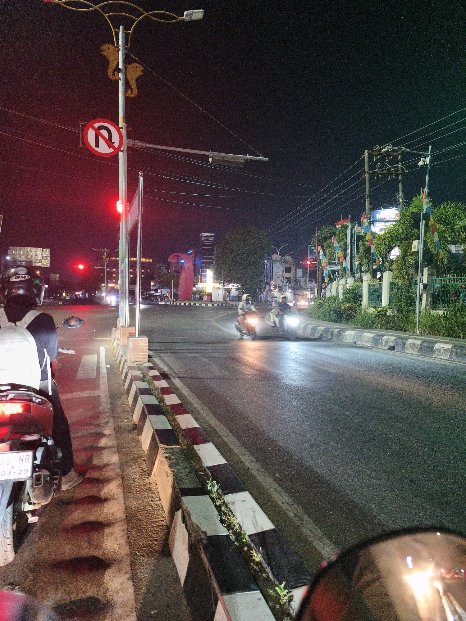 The image depicts a night scene in Samarinda, Indonesia, at an intersection. A traffic light is visible, displaying a red signal. A "No U-Turn" sign is prominently displayed on a street light pole. The road surface appears wet, suggesting recent rain. Several motorcycles are on the road, some in motion, their headlights illuminating the scene. One motorcycle, with a rider wearing a helmet and backpack, is stopped at the intersection, facing away from the camera. Buildings and palm trees line the street, and various flags are flying from poles. The overall atmosphere is that of a typical urban intersection at night, with the presence of vehicles, traffic control signals, and roadside infrastructure.