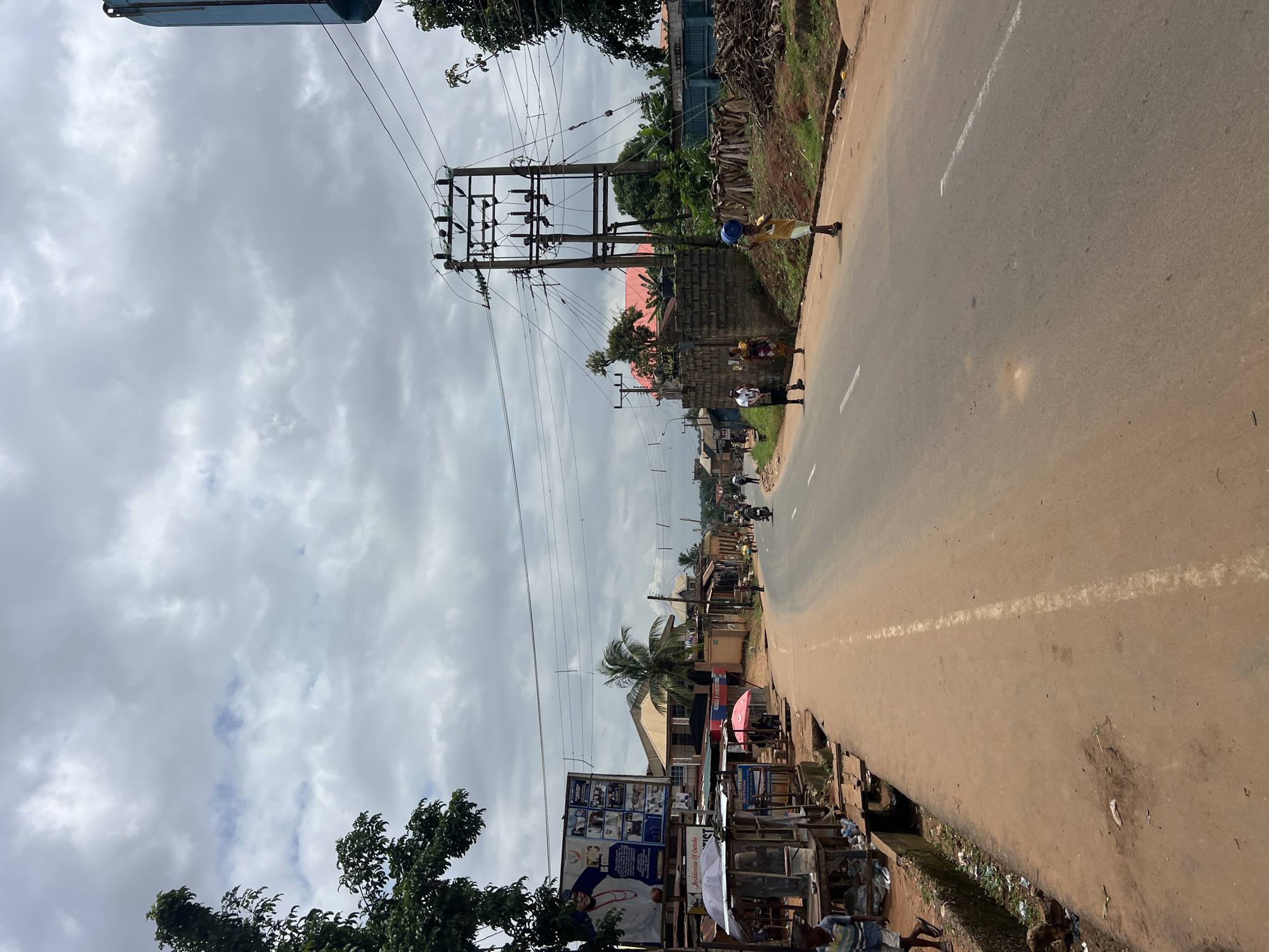 The image depicts a street scene in Nkpor, Nigeria, on an overcast day. The road, paved with asphalt and marked with white lines, curves gently into the distance. To the left of the road, a dirt shoulder is lined with makeshift stalls, some covered by tarps, selling various goods. A large billboard featuring a man in traditional attire and smaller images is prominently displayed. Further along this side, a few individuals are present, and debris is visible on the ground.
On the right side of the road, a boy carries a blue bucket on his head. Several other people are walking along the sidewalk or crossing the street. A large electrical transformer with numerous wires is situated on this side, with a stack of firewood nearby. Beyond the transformer, buildings with corrugated metal roofs and some vegetation are visible. Trees are present on both sides of the road, with one large tree on the left in the foreground. Power lines crisscross the sky, supported by poles. The overall impression is that of a busy, everyday street in a Nigerian town.