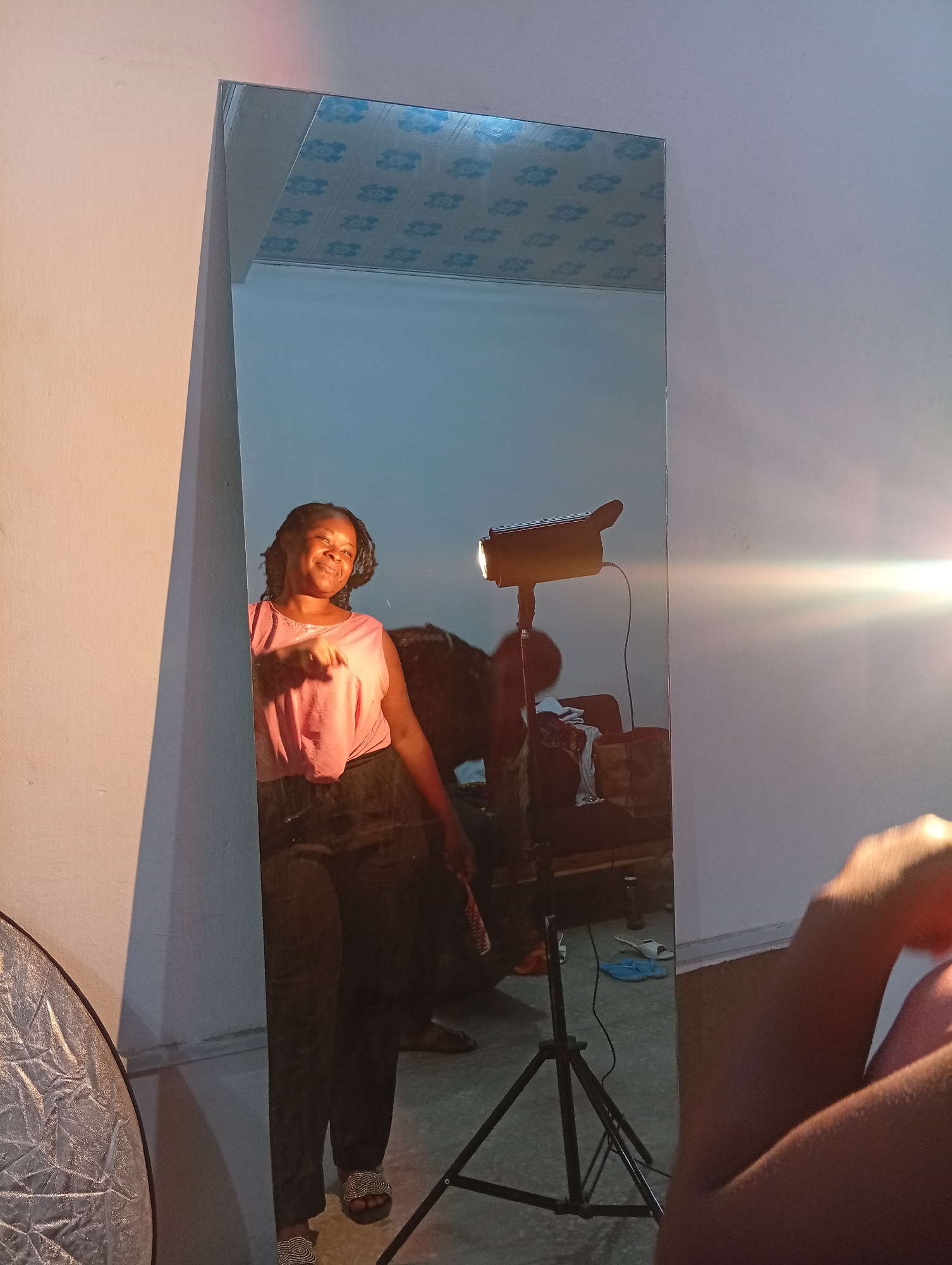 A full-length mirror reflects a woman standing in what appears to be a room. The woman is wearing a pink tank top and dark pants, with beaded sandals on her feet. She is smiling and gesturing with her right hand. To her left, a light reflector is visible. In the background of the mirror's reflection, there is a studio light on a stand, some furniture, and what looks like a patterned ceiling. The lighting in the room is warm, with a strong light source creating a lens flare to the right of the image. The environment is indistinct due to the reflective nature of the mirror and the lighting conditions. The woman is the primary subject, captured in a candid moment within a studio-like setting.