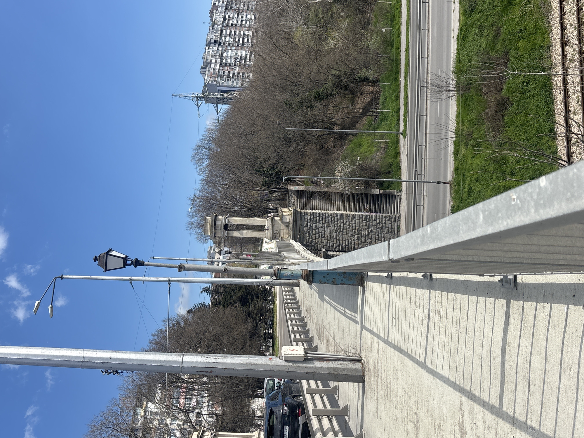 The image captures a perspective from a bridge in Varna, Bulgaria, looking towards an intersection with a set of stairs leading down to a lower level. The bridge features metal railings on the right side, and evenly spaced light poles running along its length. A dark-colored car is parked on the left side of the bridge. Below the bridge, a two-lane road is visible, bordered by grassy areas and bare trees. Beyond the trees, a tall residential building stands in the distance against a clear blue sky. Power lines are visible above the trees and the bridge.