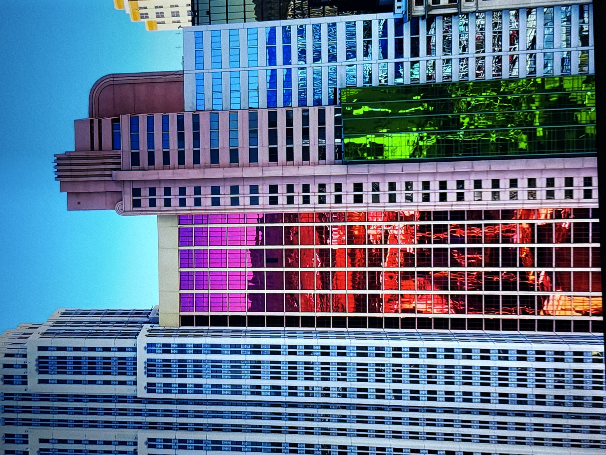 The image displays a close-up view of several modern skyscrapers against a clear blue sky. The buildings are characterized by their distinct architectural styles and colored glass facades.
On the far left, a tall, white skyscraper with numerous narrow, blue-tinted windows is visible. Its design features vertical striations and setbacks at different levels, giving it a classic, Art Deco-inspired appearance.
To the right of the white skyscraper is a building with a facade composed of intensely colored glass, predominantly vibrant pink and red. This glass reflects the surrounding environment, creating a mosaic of light and color across its numerous windows. The reflections suggest the presence of other structures or the sky.
Further to the right, a lighter-colored building with a more subdued, pinkish hue features a series of dark windows. Above this, a portion of a reddish-brown skyscraper with a distinctive stepped top and rounded corner is visible.
Next to the pinkish building is a skyscraper with a facade of light blue glass, reflecting the sky and other elements of the urban landscape.
Finally, on the right side of the image, a section of a building with green glass is seen, offering a striking contrast to the adjacent structures.
The overall scene depicts a segment of a dense urban environment, characterized by modern high-rise architecture. The focus is on the interplay of light and color on the glass surfaces of the buildings, with no people or any discernible actions or interactions depicted. The environment is clearly urban, with a clear blue sky overhead. This image does not provide any information that would allow for the identification of a specific location in Burgas, Bulgaria.