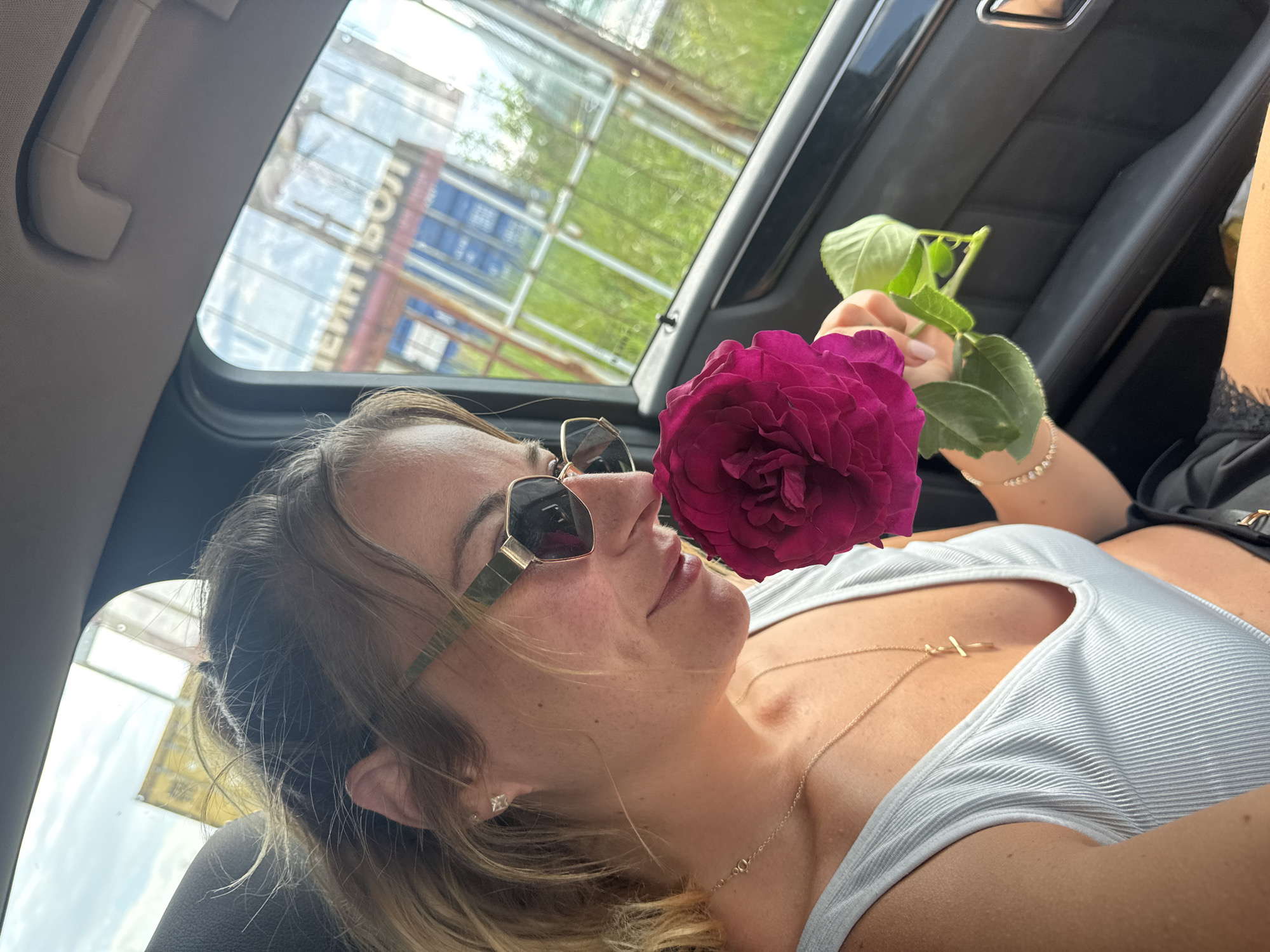 A person with sunglasses is lying in a car holding a large, magenta-colored flower with green leaves near their face. The person is wearing a white top and a gold necklace, and a diamond earring is visible in their ear. Outside the car window, a building with Russian lettering is visible, and green trees are also present.