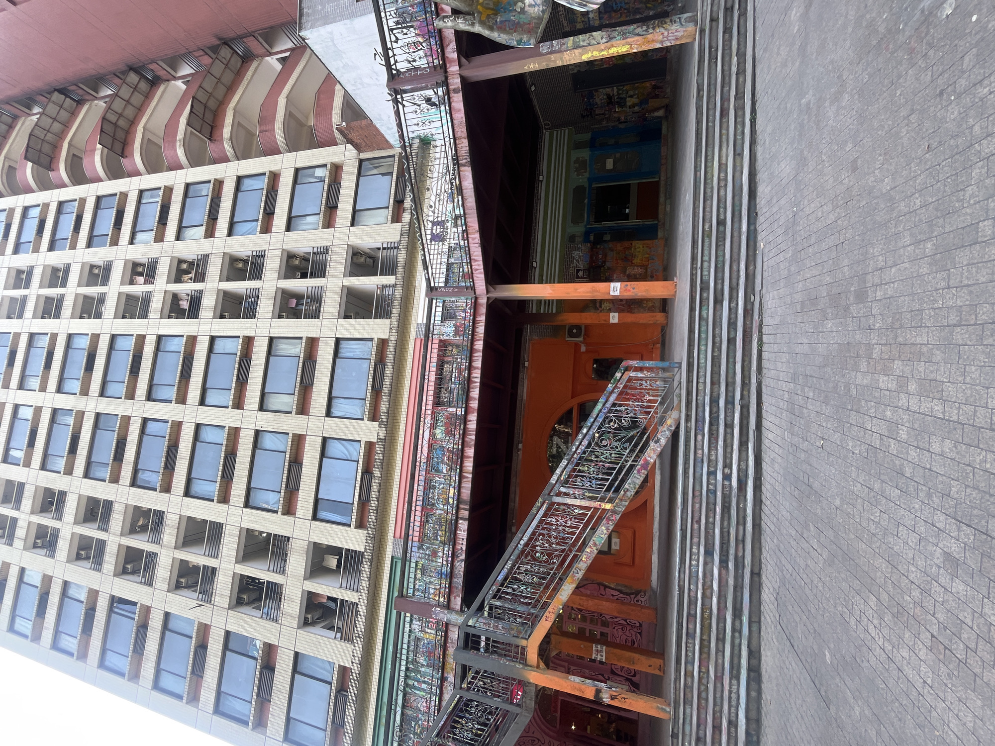 The image depicts an outdoor urban scene in Jiulongpo District, China. An ornate metal staircase covered in graffiti descends from a multi-story building with a red and white upper section and many windows, ending near a brick sidewalk. An orange doorway is visible at the bottom of the staircase, leading into a structure also adorned with graffiti.