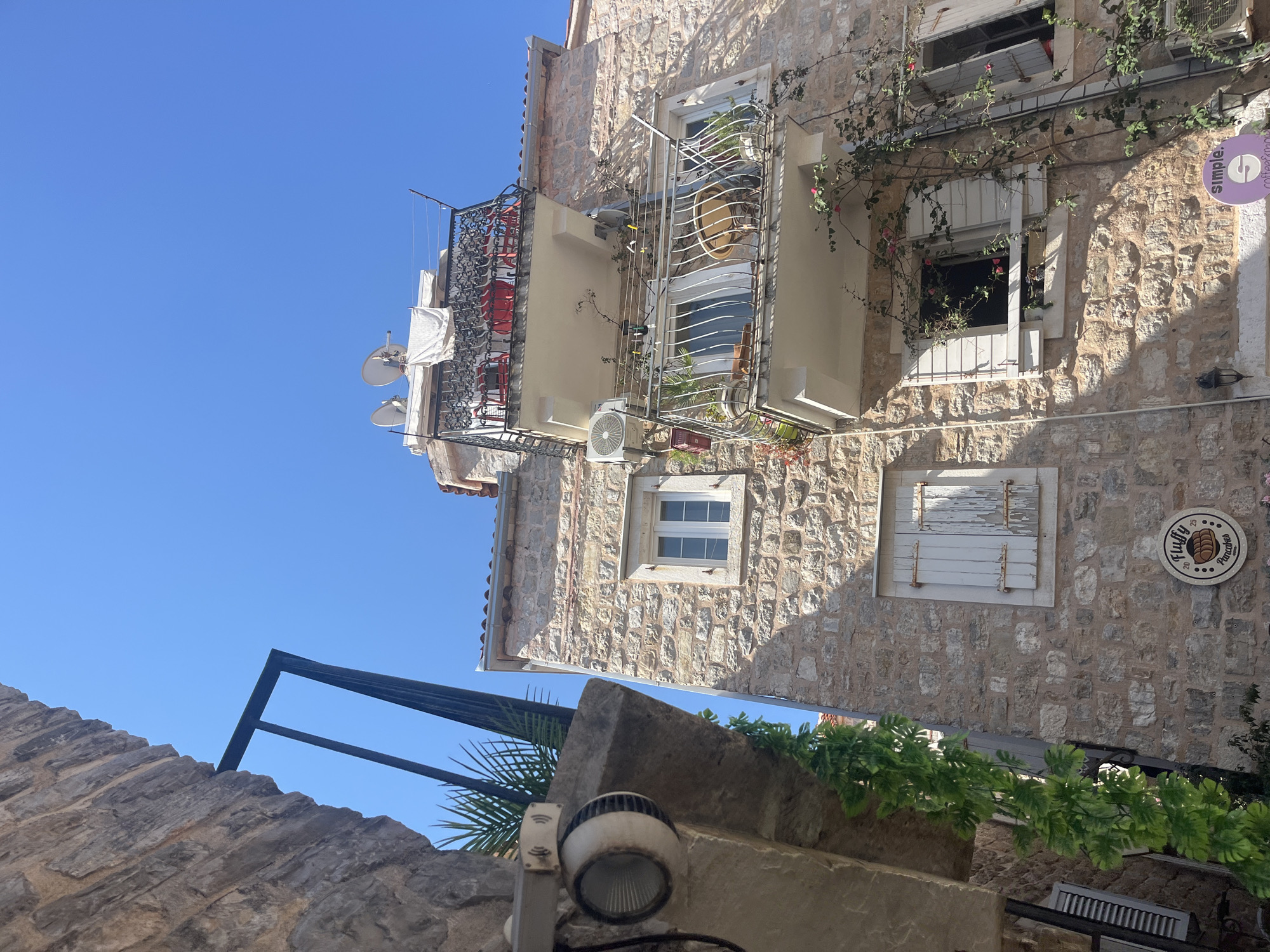 This is a low-angle, vertical outdoor shot of a stone building facade on a clear day. The building is old, with rough-hewn stones forming its walls. There are multiple windows and balconies adorned with metal railings and climbing plants with small pink flowers.
On one balcony, white laundry hangs from a railing. Another balcony has a decorative metal grille with red fabric visible behind it. Below the windows, modern air conditioning units are visible. A circular purple sign with white text "simple. coffee & snacks" is attached to the wall near a window. Another circular sign with a graphic of loaves of bread and text "Fluffy Pancakes" is also present.
The scene suggests a sunny day, likely in a Mediterranean location given the stone architecture and vegetation. The clear blue sky dominates the upper portion of the image. A black metal structure, possibly a railing or part of a roof, juts in from the left side of the frame. In the foreground, a section of a stone wall and a spotlight are visible, indicating a street-level perspective. The overall impression is one of an old European town, possibly in a tourist area given the cafe sign. There are no people visible in the image.
