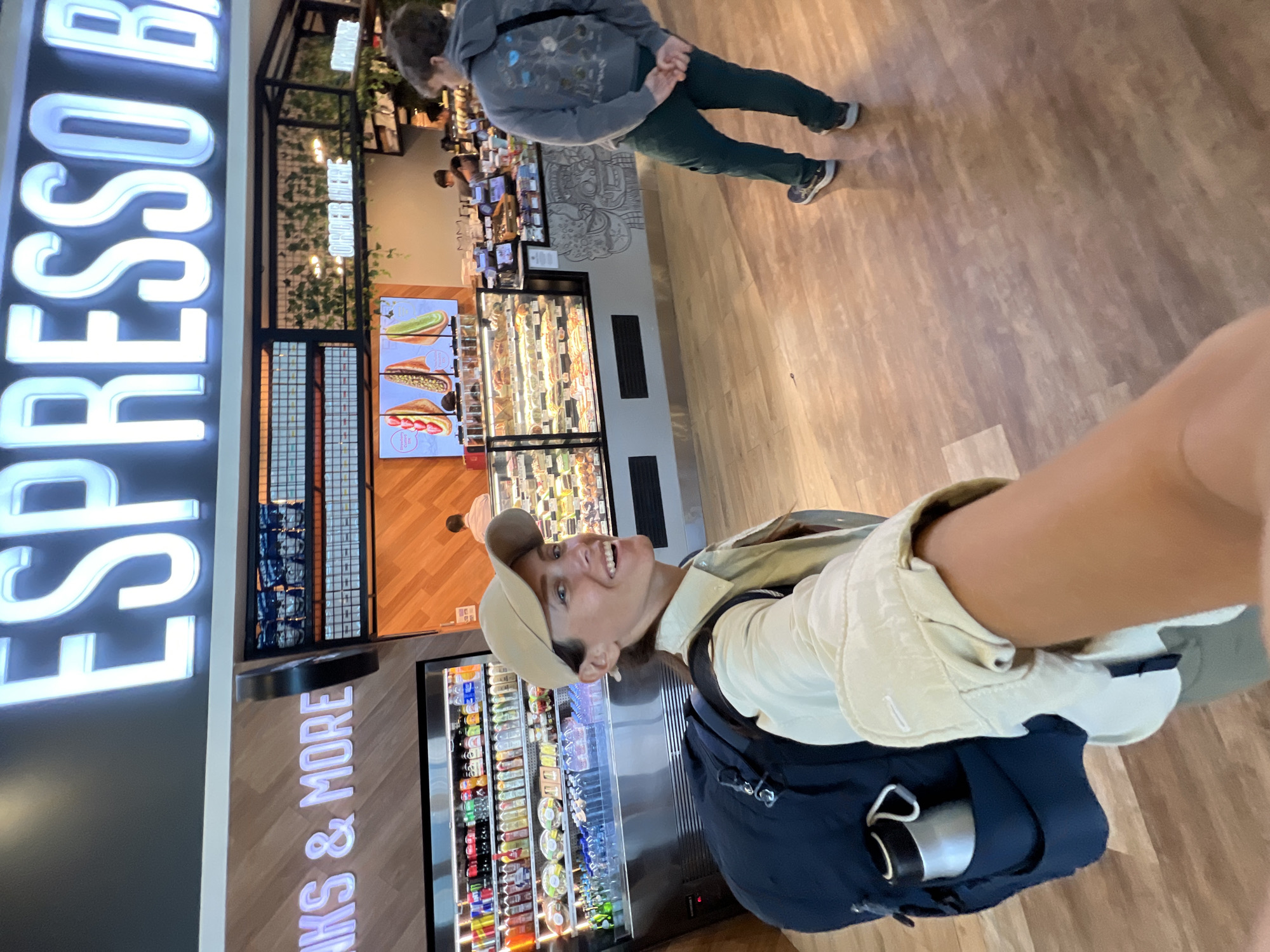 A woman wearing a tan baseball cap and a light-colored button-up shirt is taking a selfie in what appears to be an airport or a large cafe in Kuta, Indonesia. She has a large dark blue backpack on her left shoulder, with a silver water bottle visible in a side pocket. The woman is smiling broadly.
Behind her, a refrigerated display case is filled with various drinks. To her right, a counter with food displays is visible, with a menu screen showing pictures of what seem to be sandwiches or wraps. A sign above the counter reads "ESPRESSO B". In the background, another person, possibly a male wearing a grey hoodie and dark pants, is walking away from the camera. The environment has wooden flooring and wood-paneled walls. The lighting is bright, indicating an indoor commercial space.