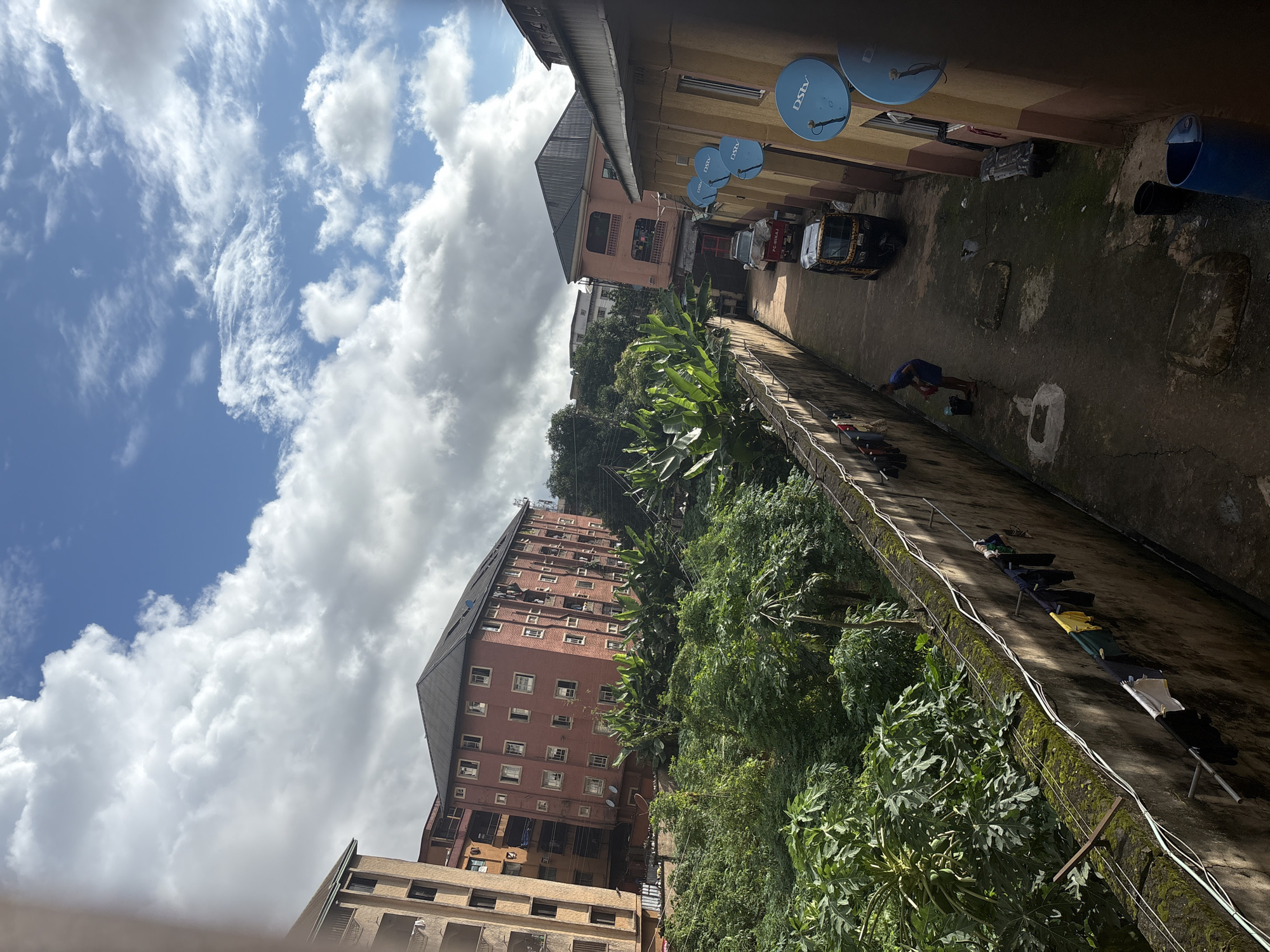The image depicts a view from a slightly elevated position looking down a narrow alleyway in Onitsha, Nigeria. The alley is bordered by a tall, weathered concrete wall on the left, on which laundry hangs on a clothesline. Lush green vegetation, including what appear to be banana trees, grows densely behind the wall. Across the alley, a yellow and brown building features several satellite dishes mounted on its exterior. Further down the alley, a yellow auto-rickshaw is parked. A person, dressed in a blue shirt and shorts, is visible on the right side of the alley, appearing to be engaged in some activity near a bucket. The sky above is a mix of blue and white clouds. The overall impression is of a residential area with vibrant plant life and typical urban elements of a Nigerian city.