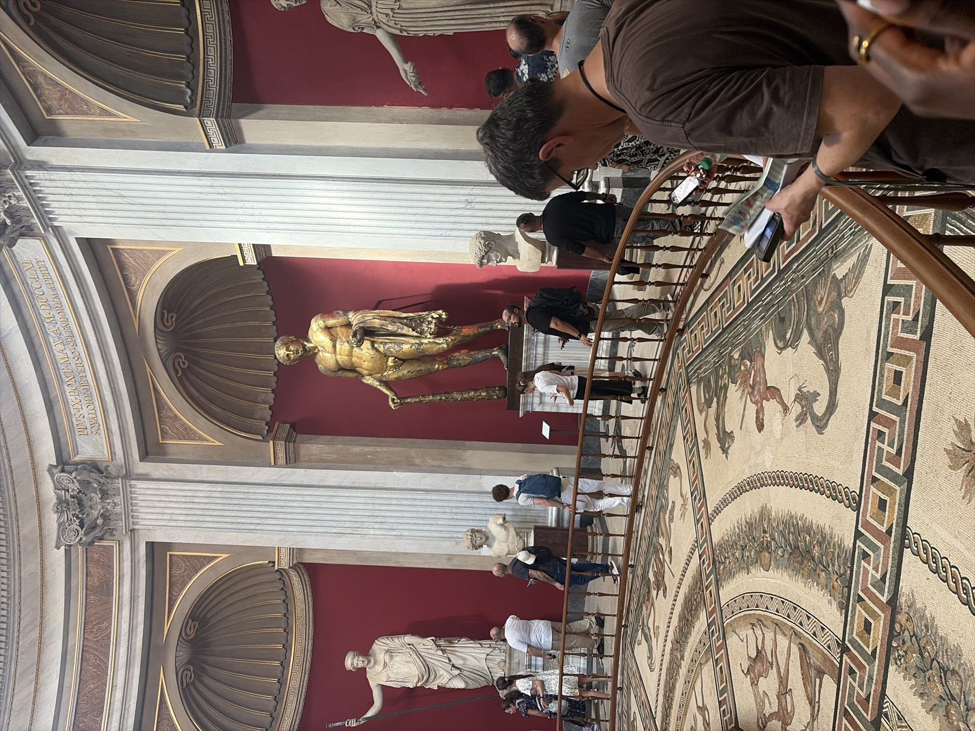 This image depicts the interior of a museum in Rome, Italy, likely the Pio Clementino Museum within the Vatican Museums. The scene is an exhibit featuring ancient Roman sculptures and mosaics. In the center, a large, gilded bronze statue of Hercules, identified as the "Hercules Farnese," dominates the space, holding a club. To the left, a tall white marble statue of a female figure, possibly Minerva, stands with a spear. To the right, a partial view of another large marble statue is visible. Several busts are also displayed, including one of Emperor Augustus.
The floor is covered with an intricate mosaic featuring depictions of mythological scenes with figures and animals. A group of tourists is present, observing the artworks. One individual in the foreground, a man wearing a brown t-shirt, is holding a smartphone and looking at a guidebook or brochure. Other visitors are seen standing and walking along a curved wooden railing that separates them from the exhibit. The walls are a deep red, and architectural elements like fluted columns and arched niches are visible. The lighting suggests an indoor environment, likely illuminated by artificial lights supplemented by natural light from high windows or skylights.