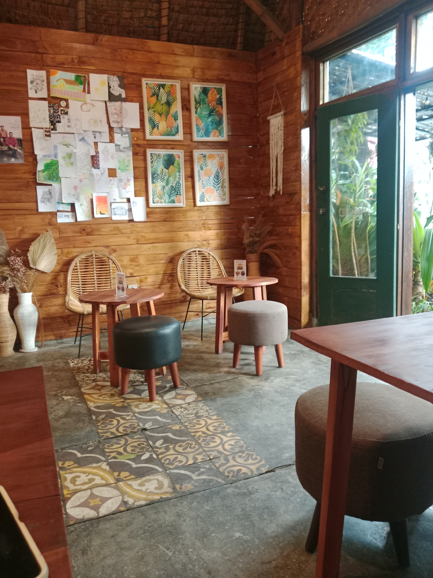 The image shows the interior of a cafe with wooden walls and a thatched roof. In the foreground, a dark wooden table with a reddish-brown surface is on the right side of the frame, with a round, fabric-covered stool with dark wooden legs beneath it. A tiled rug with a geometric pattern in yellow, gray, and white is on the floor between two small, round wooden tables. Each of these tables has a single dark stool or ottoman with wooden legs. Behind these tables, two rattan chairs are visible. The wooden wall is decorated with numerous drawings and framed botanical prints. To the right, a green-framed glass door and window provide a view of lush green plants outside. Two decorative vases, one tall and white and the other shorter and brown, are placed on the floor near a dried plant arrangement on the left side of the image. This establishment is likely a cafe in Bandung City, Indonesia, given the tropical greenery visible through the glass and the prevalent use of natural materials like wood and rattan, common in Southeast Asian cafe designs.