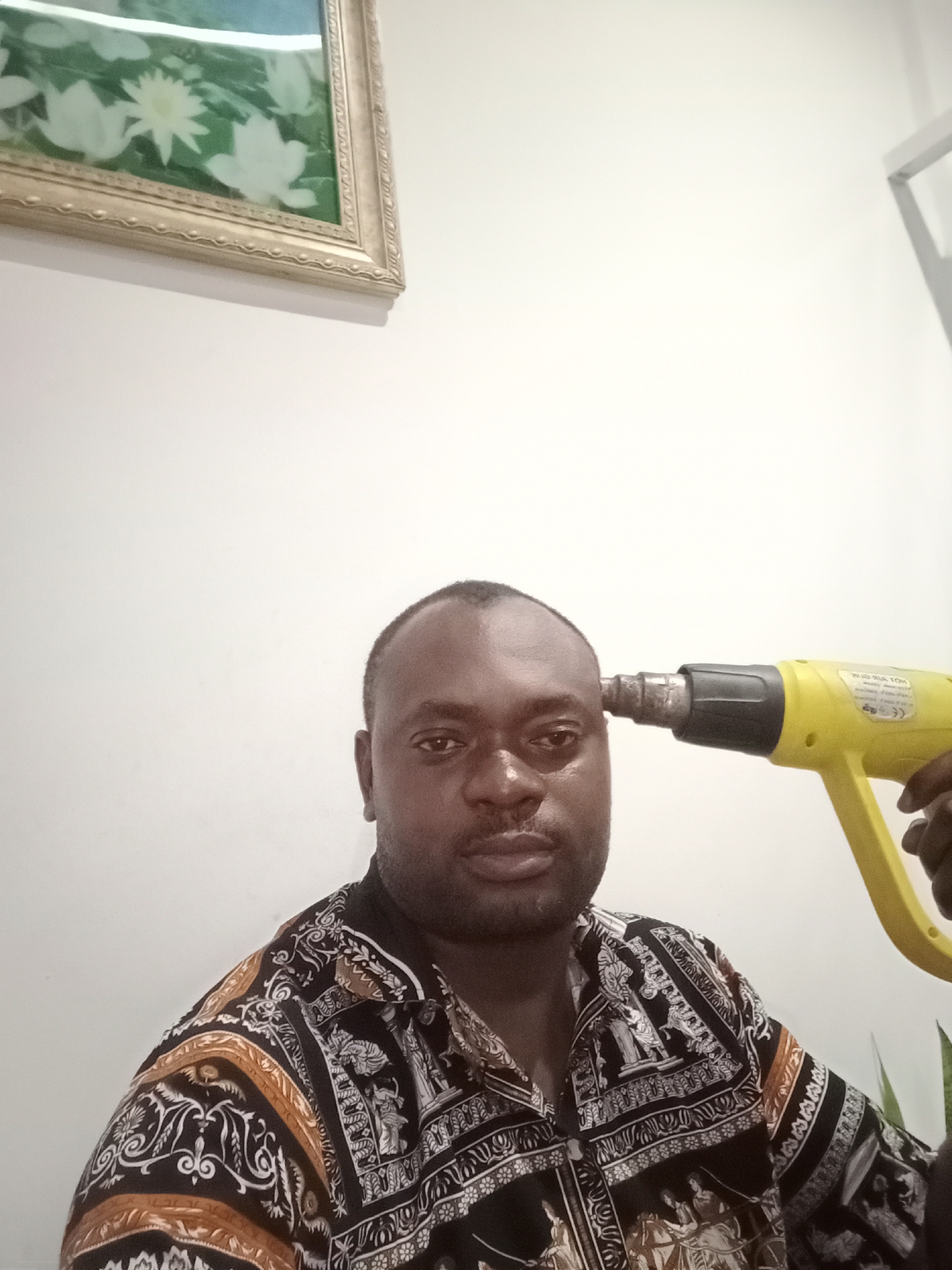 A dark-skinned man is holding a yellow heat gun to his head. He is wearing a patterned shirt. In the background, there is a painting on the wall and a partially visible white frame. It is not possible to confirm the location as Gwarinpa, Nigeria, based solely on the image.