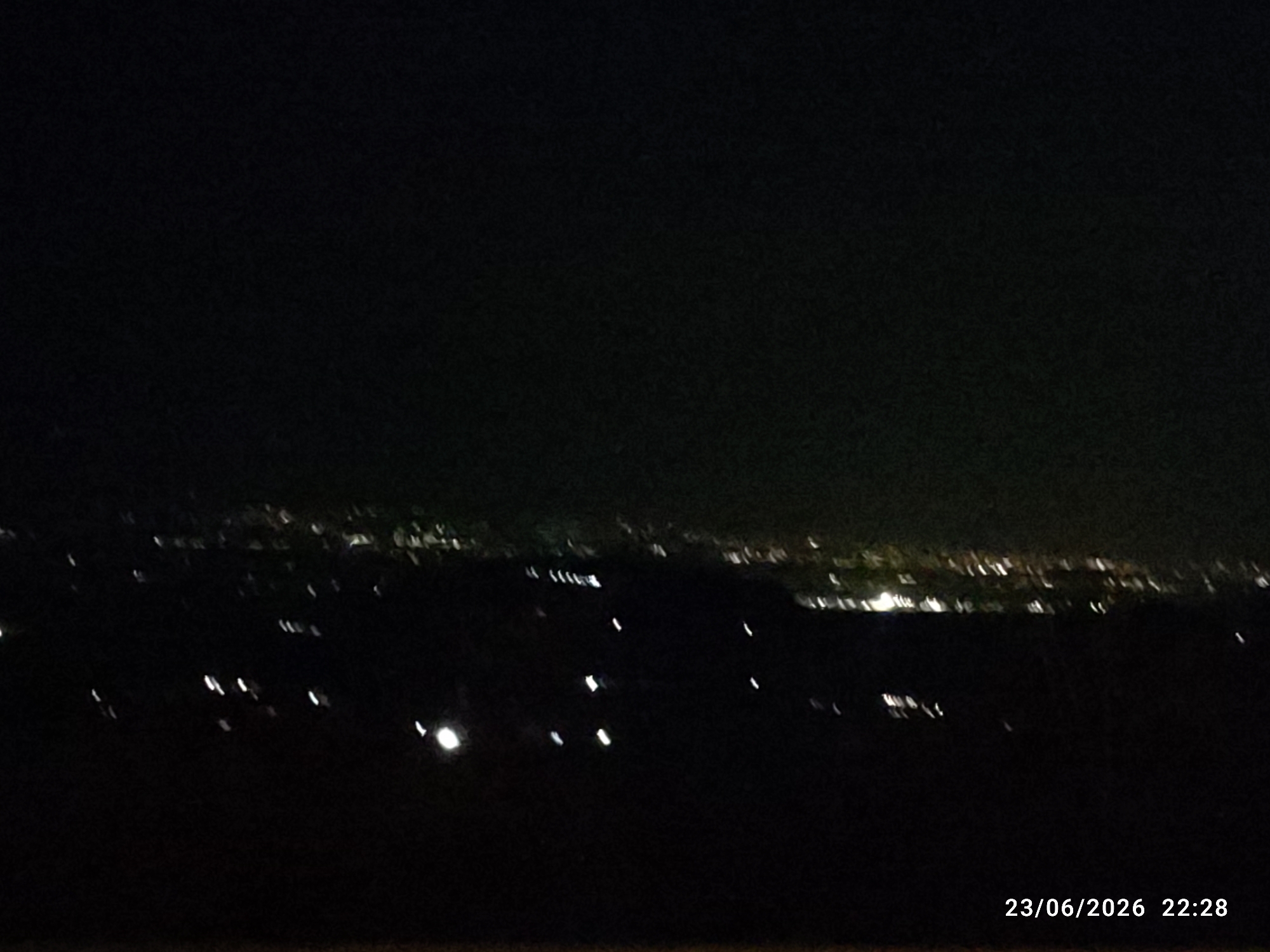 This is a nighttime, landscape photograph taken from an elevated position overlooking a city or town. The foreground is dark and indistinct, suggesting it is a natural landscape such as hills or fields. In the midground and background, numerous lights are visible, indicating urban development. These lights appear as blurred streaks and dots, characteristic of a long exposure or the limitations of low-light photography. The lights form a horizon line across the image, suggesting a settlement spread out below. Some clusters of lights appear more concentrated, possibly representing the denser areas of a town. The overall scene depicts a view of human habitation at night.
A timestamp is visible in the lower right corner of the image, reading "23/06/2026 22:28". This indicates the photograph was taken on June 23, 2026, at 10:28 PM. The time of day is clearly night, and the dark sky supports this. There are no discernible weather conditions, but the darkness suggests a clear or at least not overcast night. No people or specific objects are clearly identifiable. The location context of Scordia, Italy, would suggest this is a view of that town or its surroundings. The mood is quiet and serene, evoked by the darkness punctuated by the distant glow of lights.