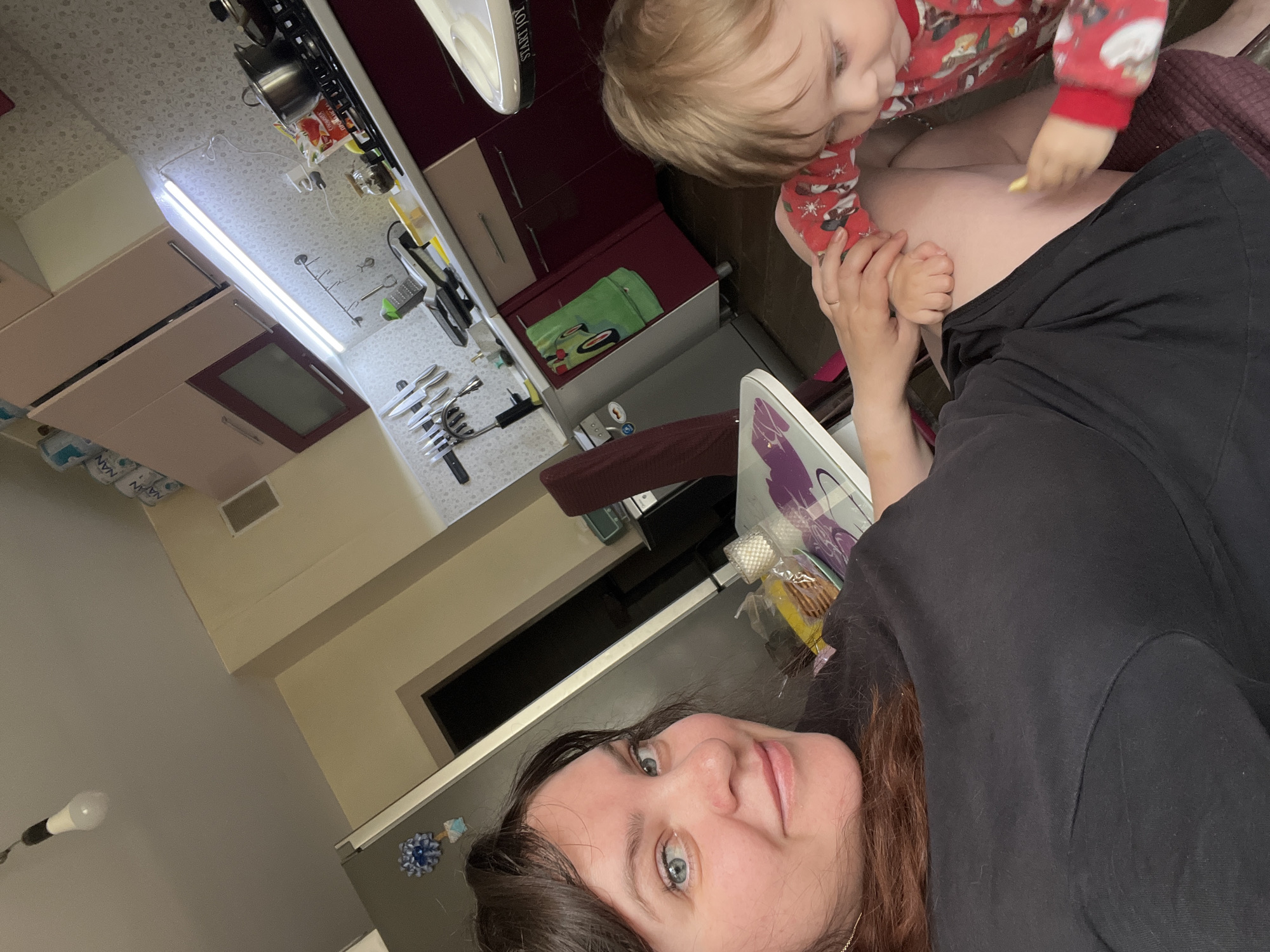A candid, upright, portrait-oriented image captures an adult woman and a young child in a home kitchen setting in Noviy, Russia. The woman, in the foreground, has long dark brown hair and blue eyes, looking directly at the camera with a neutral expression. She wears a black top. Perched on her lap or hip is a young child with blonde hair, dressed in a red, patterned long-sleeved top. The child is focused downwards, holding a small yellow object on the woman's thigh.
The kitchen background reveals a functional space. A countertop runs horizontally behind the subjects, laden with various kitchen items including a knife block filled with knives, a grater, and other utensils. A stovetop with a pot is visible further back. Above the counter, light-colored upper cabinets are interspersed with darker, possibly maroon-toned cabinets. A long, bright fluorescent light fixture illuminates the workspace. To the left, a beige wall features a single hanging light bulb.
Visible details include a white appliance with partially discernible text "STARTIOV" or similar, a green container with a car design, and a red and white package with Cyrillic text, likely a food item. In the upper left, partially visible blue packaging bears the text "NAN". A refrigerator with a blue flower-shaped magnet is also present behind the woman's head. The scene suggests a relaxed, everyday domestic moment, likely indoors during daytime or evening given the artificial lighting.