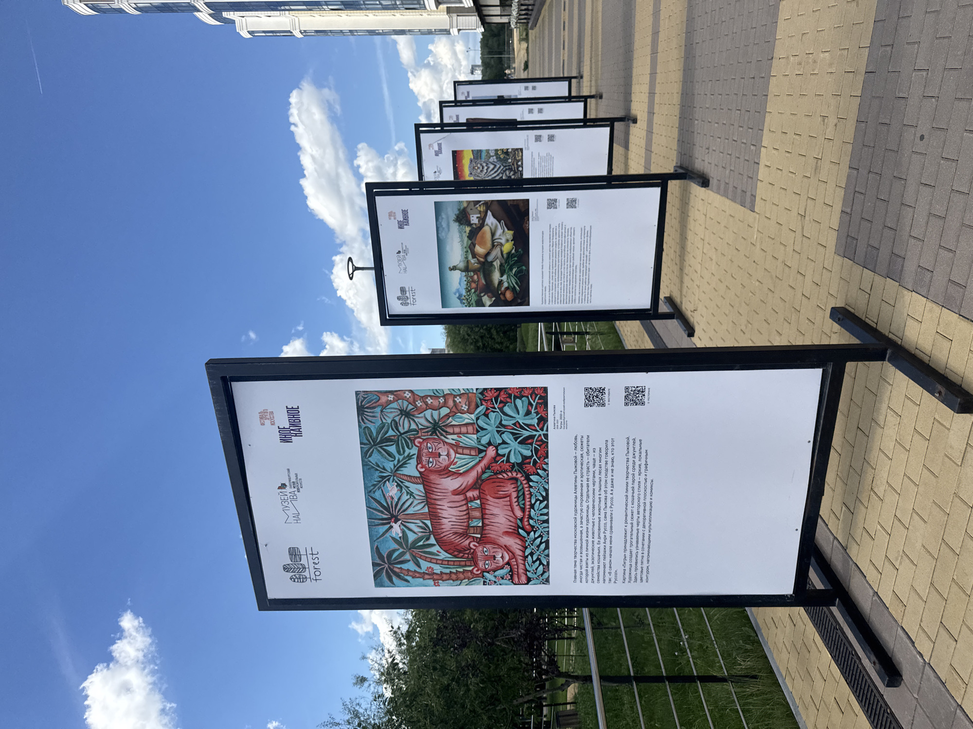 This is an outdoor exhibition of paintings, with multiple display boards arranged in a line on a paved walkway. The first board on the left features a painting of two tigers against a jungle-like background. The text on this board describes the artwork as "Tigers" by Alevtina Pyshkova, created in 2010, and mentions it is an "exhibition of folk art." The other boards in the background also display paintings and accompanying text, though they are less clear. In the background, there is a green lawn with trees on the left and a modern residential building with balconies on the right. The sky is blue with scattered white clouds. The scene appears to be in a public space, possibly a park or a pedestrian area, in Yekaterinburg, Russia, given the text and the presence of outdoor exhibitions. There are no people visible in the immediate foreground or interacting with the exhibition.