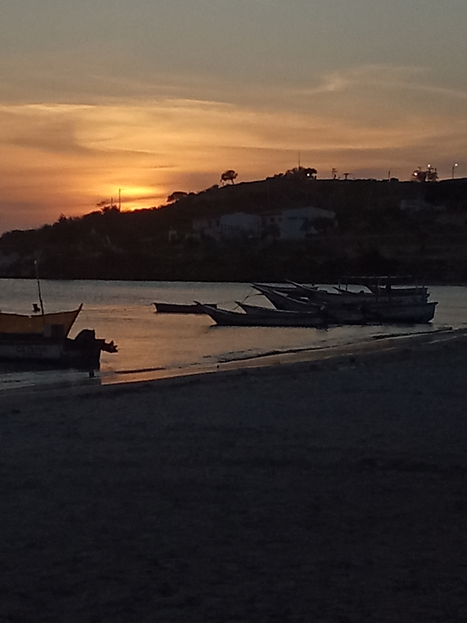 The image captures a sunset scene in Juan Griego, Venezuela. The sun is setting over a hill, casting an orange glow across the sky and water. Several small boats are docked near the shore, their silhouettes visible against the light. A beach is in the foreground. Buildings and other structures dot the top of the hill.