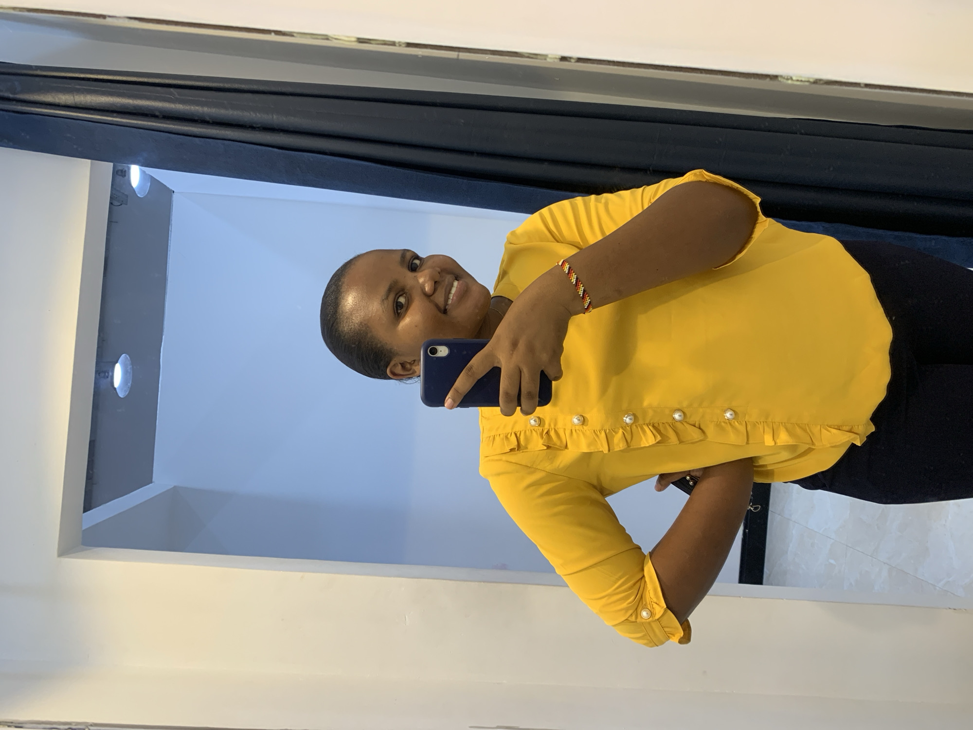 A medium shot depicts a woman holding a dark blue smartphone in her left hand, facing and smiling at the camera. She has short, dark hair and is wearing a yellow blouse with pearl button details on the sleeves and ruffled trim. A bracelet with multi-colored beads is visible on her left wrist.
She is standing in what appears to be a room with light blue or white walls that reflect the light from small circular lights placed in the corner near the ceiling. A dark blue curtain or drape is partially visible in the background. The lower half of her body is obscured, but it is evident that she is wearing a dark garment.
The location cannot be verified as being in Dar es Salaam, Tanzania without additional information.