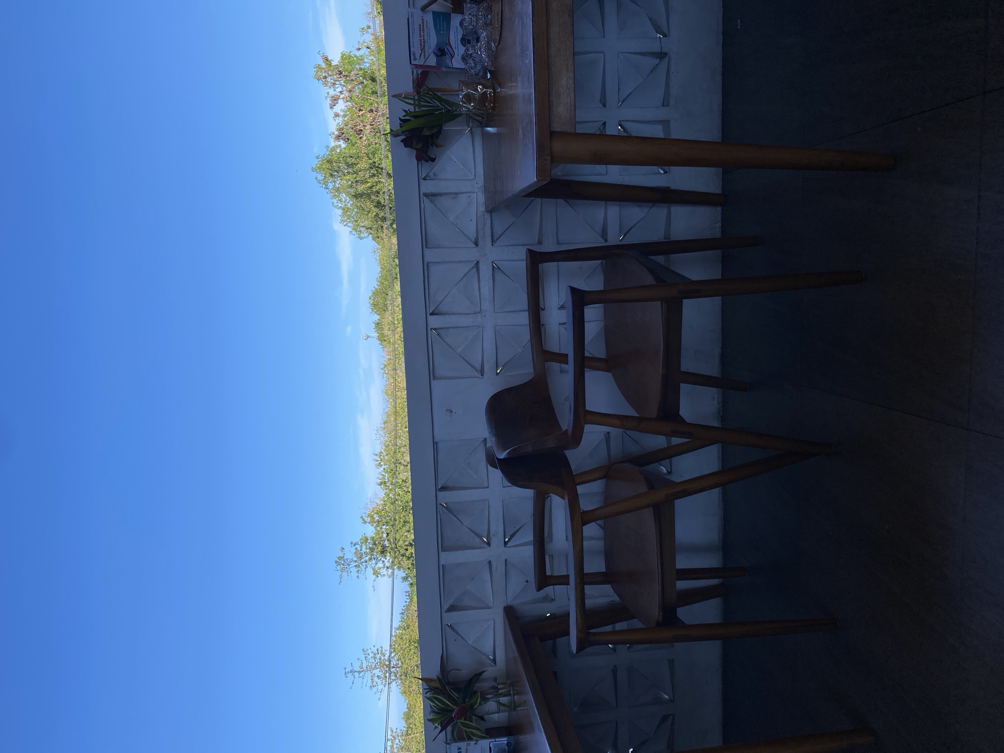 The image shows an outdoor seating area in Kuta Selatan, Indonesia, featuring wooden tables and chairs. A white wall with geometric patterns serves as a backdrop, and a line of trees and blue sky are visible in the distance. A vase with greenery and other items sit on the table. The seating area appears to be dimly lit, with shadows covering the floor. No people are visible.