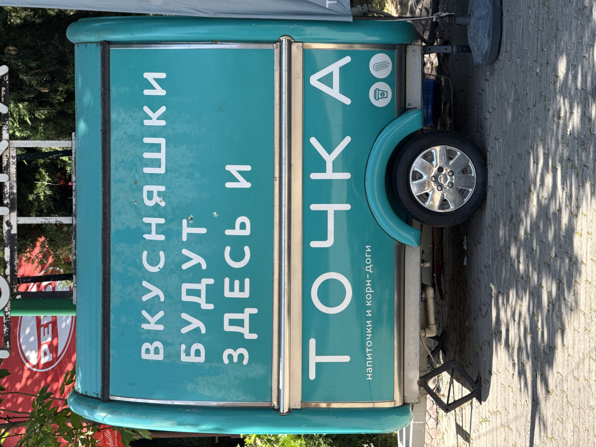 The image shows a food cart in Tashkent, Uzbekistan. The cart is turquoise with white lettering in Cyrillic. The Cyrillic text translates to "Tasty things will be here" and "Point. Drinks and corn dogs." The cart has a single wheel with a chrome rim. The ground is paved with rectangular bricks, and the cart is parked partially in the shade of a tree. There is a red and white sign visible to the left, partly obscured by foliage. A jack for lifting the cart is visible beneath the rear.