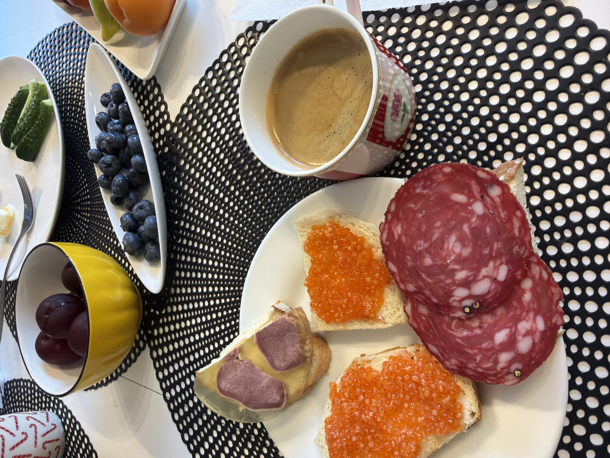In Baloži, Latvia, a table is set for a meal with a variety of food items. In the foreground, a white plate holds several appetizers. Two slices of bread are topped with vibrant orange caviar, and two circular slices of salami with white marbling are also present. Adjacent to these, another slice of bread is topped with caviar, and a piece of appetizer with what appears to be meat in aspic sits on top of bread. A pink and white patterned mug filled with dark coffee is situated to the right of the plate. In the background, a yellow bowl contains dark red grapes. A white oval dish holds a pile of fresh blueberries. Further back, another white plate displays several sliced green cucumbers. The table is covered with a black and white polka dot placemat. The setting appears to be indoors, likely a home dining area.