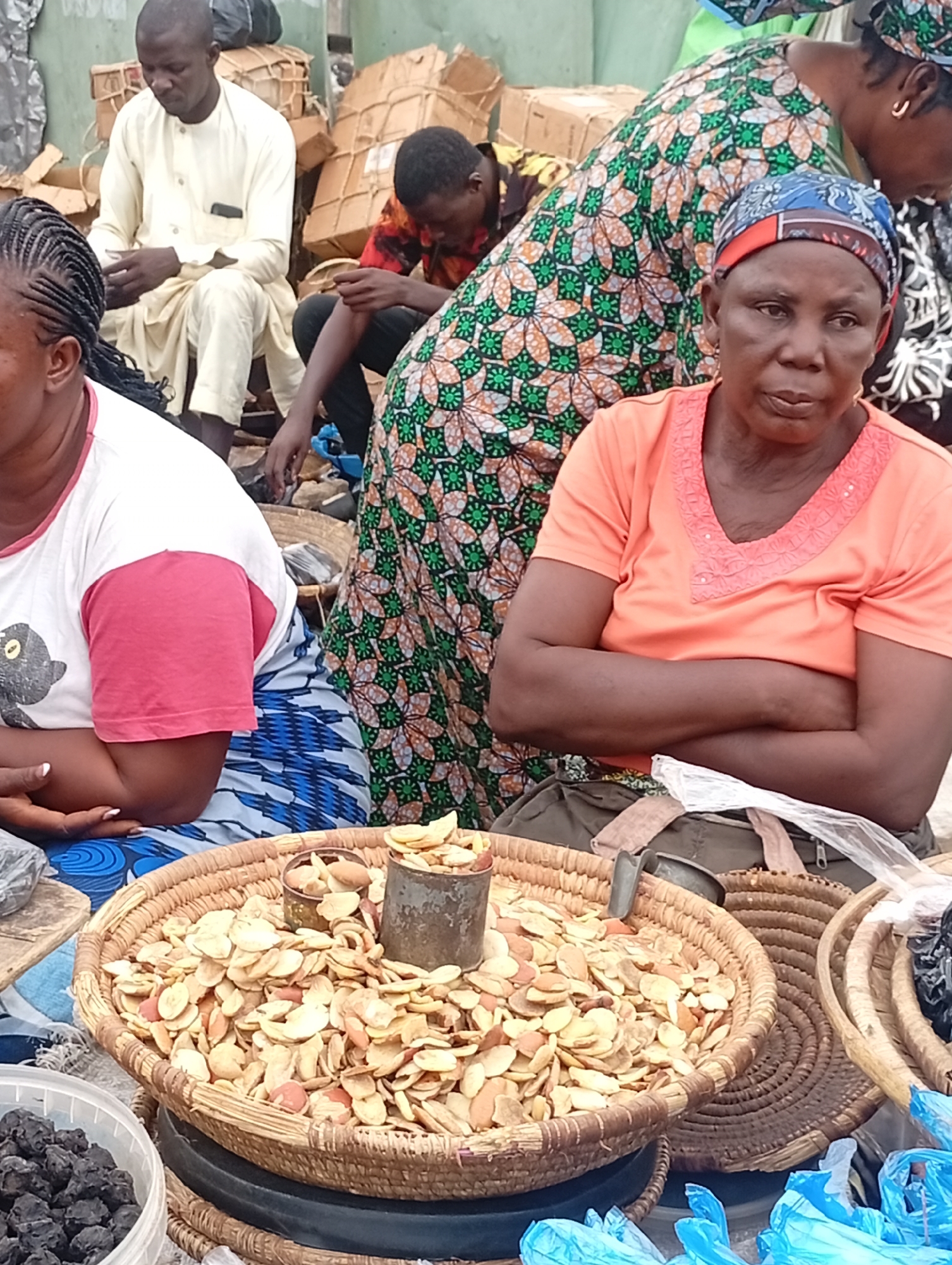 The image depicts a market scene in Maiburiji, Nigeria. A woman in an orange shirt and headscarf sits with her arms crossed, positioned in front of a large woven basket filled with what appear to be dried fruit slices, possibly nuts. Two metal containers sit atop the fruit. To her left, another woman in a white and red shirt with blue patterned fabric is partially visible. Behind them, a person in a green and orange patterned dress stands next to a man wearing a cream-colored outfit. Other figures are visible in the background. Several boxes and what appears to be scattered merchandise add to the market atmosphere. Additional baskets and a container of dark, round fruits are also visible near the front of the scene.