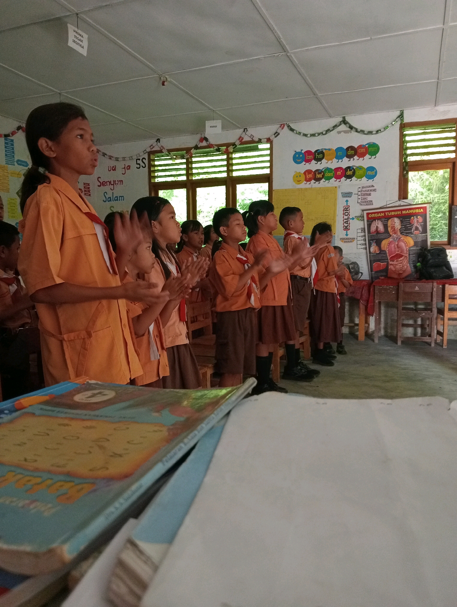 The image shows a group of elementary school children in a classroom in Balige, Indonesia. The children are wearing orange uniforms and appear to be standing and clapping. They are positioned in front of a whiteboard with educational posters and decorations. One poster displays an anatomical chart of the human body. In the foreground, several books are stacked on a desk. The classroom has large windows, suggesting natural light. The overall scene depicts a typical school day activity.