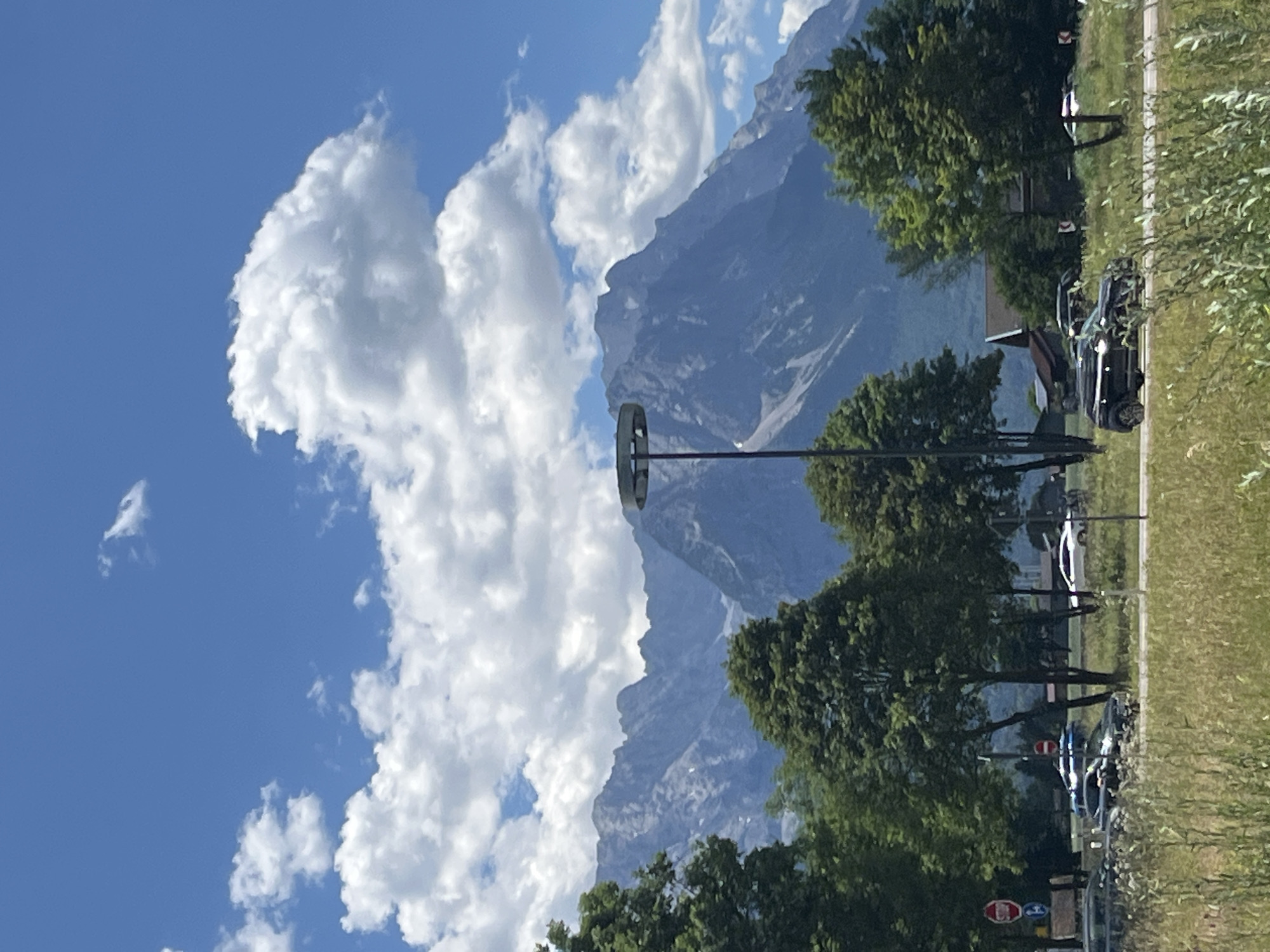 The image shows a mountainous landscape in Garmisch-Partenkirchen, Germany, under a bright blue sky with scattered white clouds. In the foreground, a road with several cars is visible, with trees and grassy fields on either side. A tall, modern street lamp with a circular light fixture stands prominently in the middle ground, in front of the mountains. The mountains themselves are rugged and appear to be covered in some patches of snow. In the background, there are some buildings visible, suggesting the presence of a town or village. The scene depicts a typical summer day in a scenic alpine region.