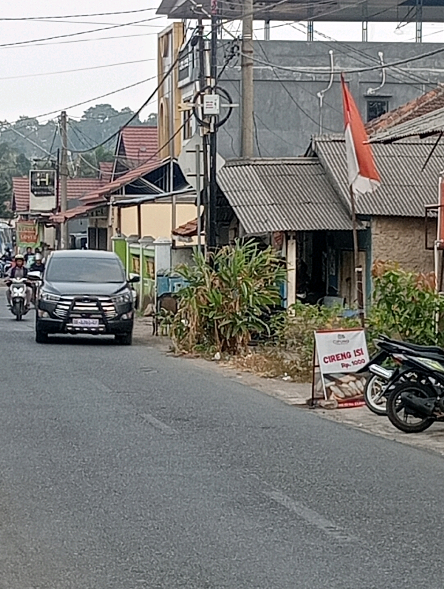 The image depicts a street scene in Bandar Lampung, Indonesia, focusing on a black SUV driving on a paved road. The SUV has a bull bar on its front. To the left of the SUV, two motorcycles are visible, with one carrying a rider. In the background, there are several buildings with red-tiled roofs and a taller building with a yellow facade. A red and white Indonesian flag is prominently displayed on a pole. A small sign on the right side of the road advertises "CIRENG ISI Rp. 1000". The environment appears to be a street with overhead electrical wires, vegetation, and other structures consistent with a residential or commercial area. The sky is overcast, and the lighting suggests it may be late afternoon or early morning.
