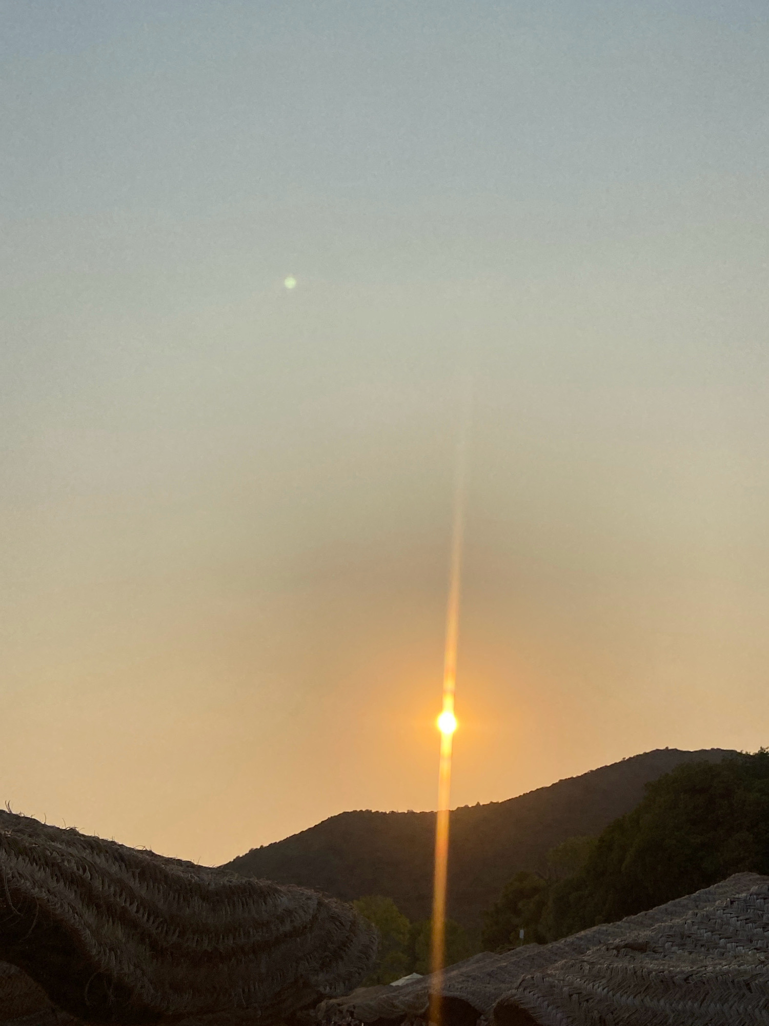 An outdoor, vertical, eye-level, medium shot shows a sunset over a hilly landscape, with the foreground occupied by thatched roofing structures. The sun, positioned slightly right of center and halfway up the frame, is a bright orb emitting a strong vertical flare. The sky transitions from a pale blue at the top to a warm orange glow near the horizon. A small, bright, greenish-white object, possibly lens flare or a celestial body, is visible in the upper left quadrant of the sky. The dark, silhouetted shapes of hills and lush green trees form the middle ground. In the foreground, two thatched roofs, appearing dry and textured, frame the bottom corners of the image. The scene evokes a tropical or coastal atmosphere, likely in a resort area. The location is specified as Prijevor, Montenegro. There are no people or animals visible in the image.