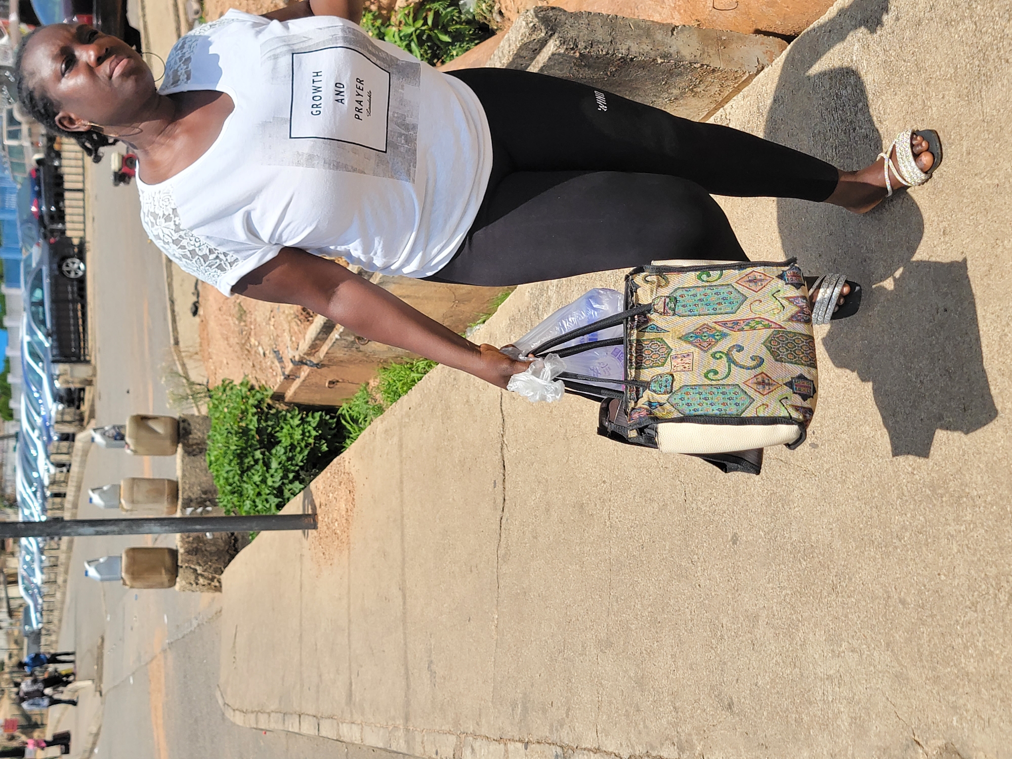 A woman is standing on a sidewalk next to a street in what appears to be Jos, Nigeria, based on the environment, style of clothing, and visual context. She wears a white t-shirt with "GROWTH AND PRAYER" printed on it, black leggings, and sandals with embellishments. She holds shopping bags and a tote bag with patterned fabric. The background shows cars parked along the street, foliage, and elements of urban infrastructure. The sidewalk appears to be made of concrete. The photograph seems to be taken during daytime.