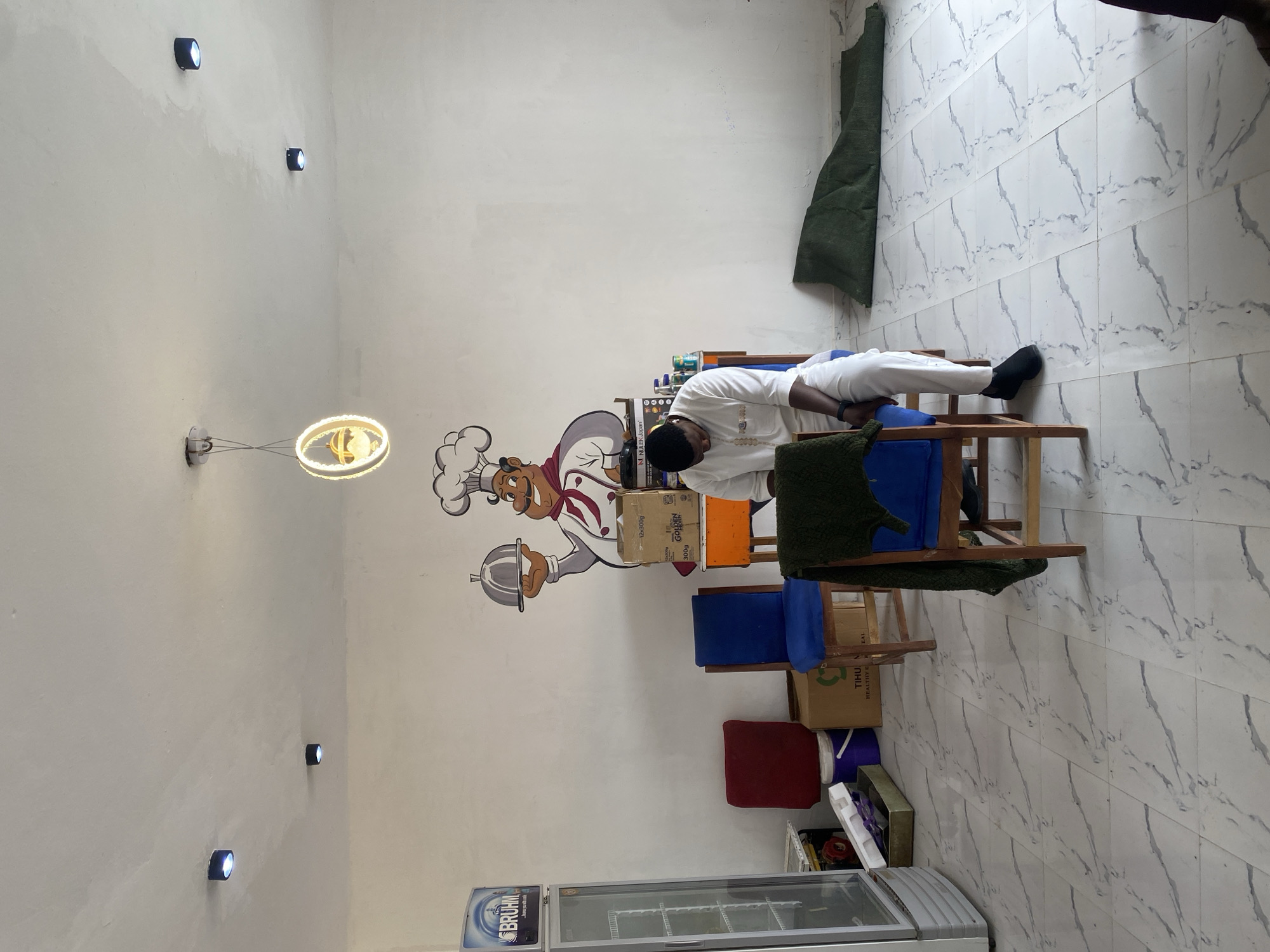 A man in a white shirt and pants is sleeping on a wooden chair in what appears to be a commercial space with tile floors and white walls. The chair is situated amidst other chairs of varying colors (blue, orange, red). A large cartoon mural of a chef is on the wall. Various boxes, containers, and other miscellaneous items are scattered around the chairs. A refrigerator is visible in the lower portion of the frame. The location is possibly in Gombe, Nigeria.