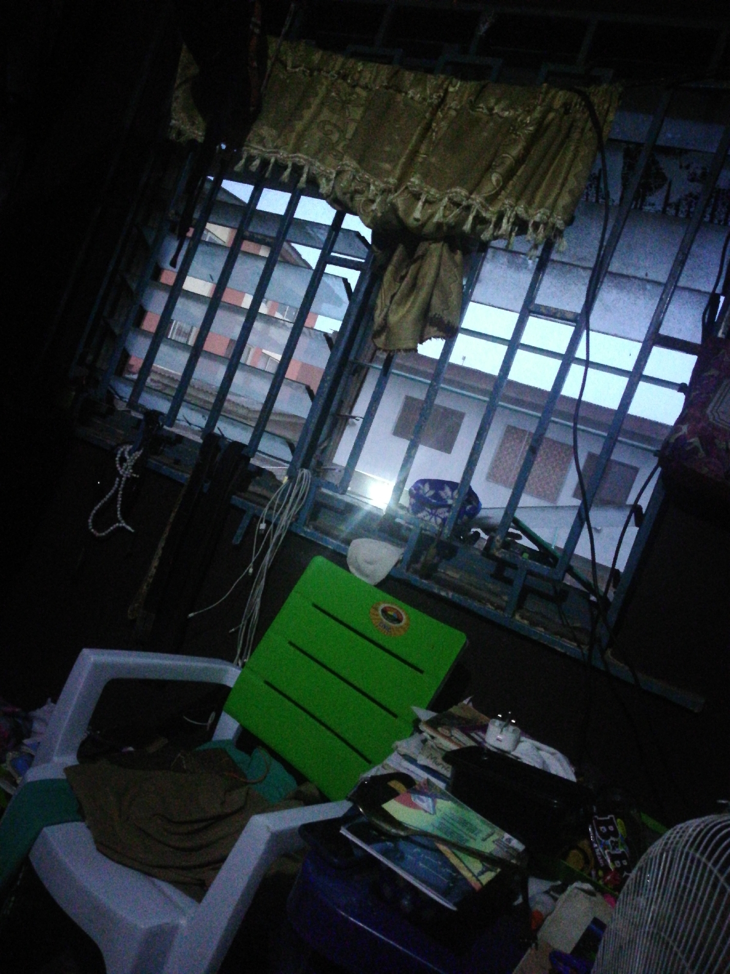 The scene appears to be inside a dark room, likely a domestic space, with a barred window as the primary source of light. Outside the window, buildings are visible, suggesting an urban environment. The window has a light-colored curtain draped over it. In the room, there's a white plastic chair with some clothing on it, a green object leaning against the window bars, and a pile of miscellaneous items including books and a fan. The overall impression is one of clutter and limited natural light.