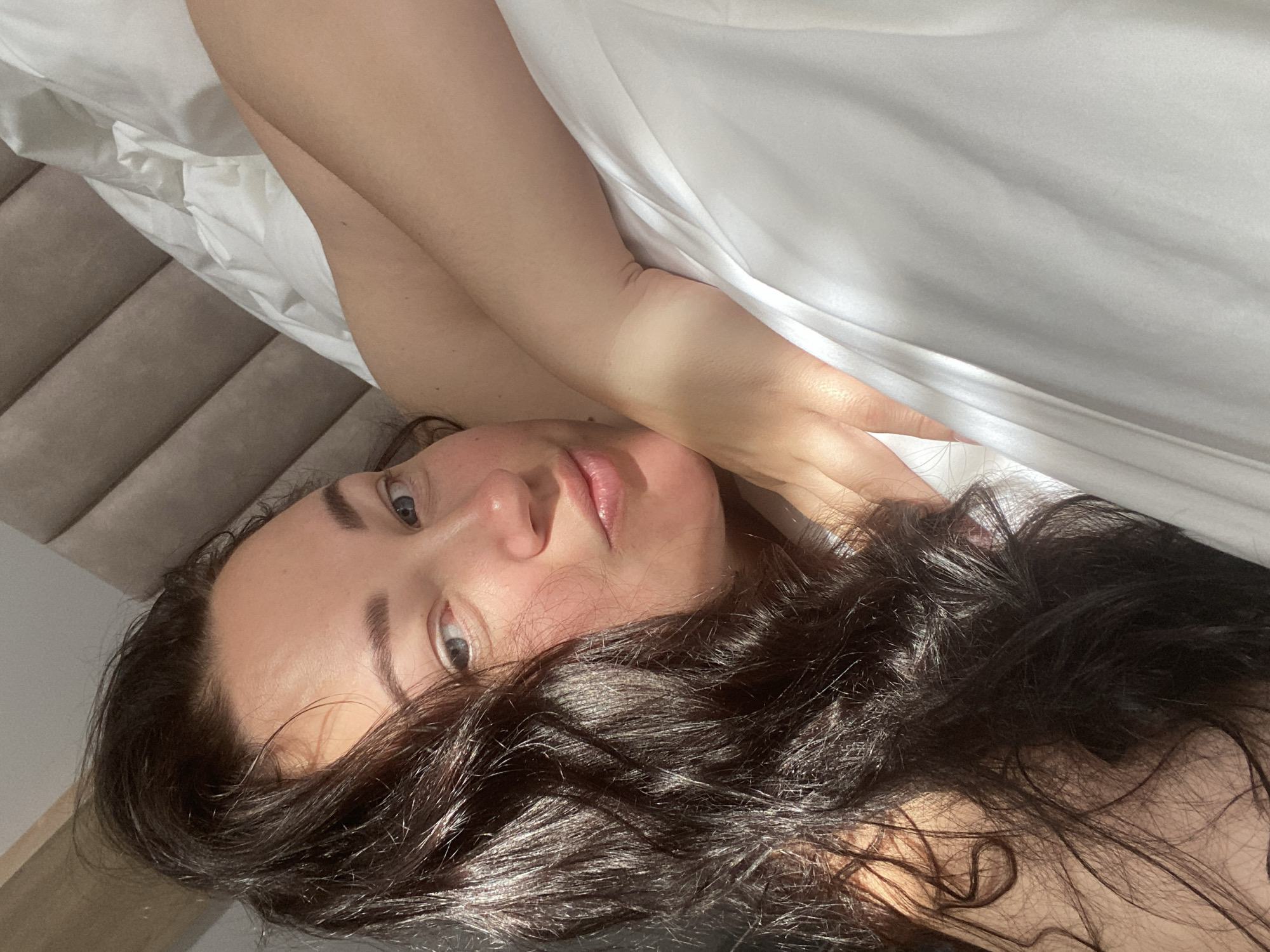 A woman lies on a bed covered with a white sheet. The bed has a gray headboard. The woman has dark hair and fair skin. She is holding her arms up near her face. The location is not discernible from the image. The city of Sochi, Russia, is not discernible from the image.