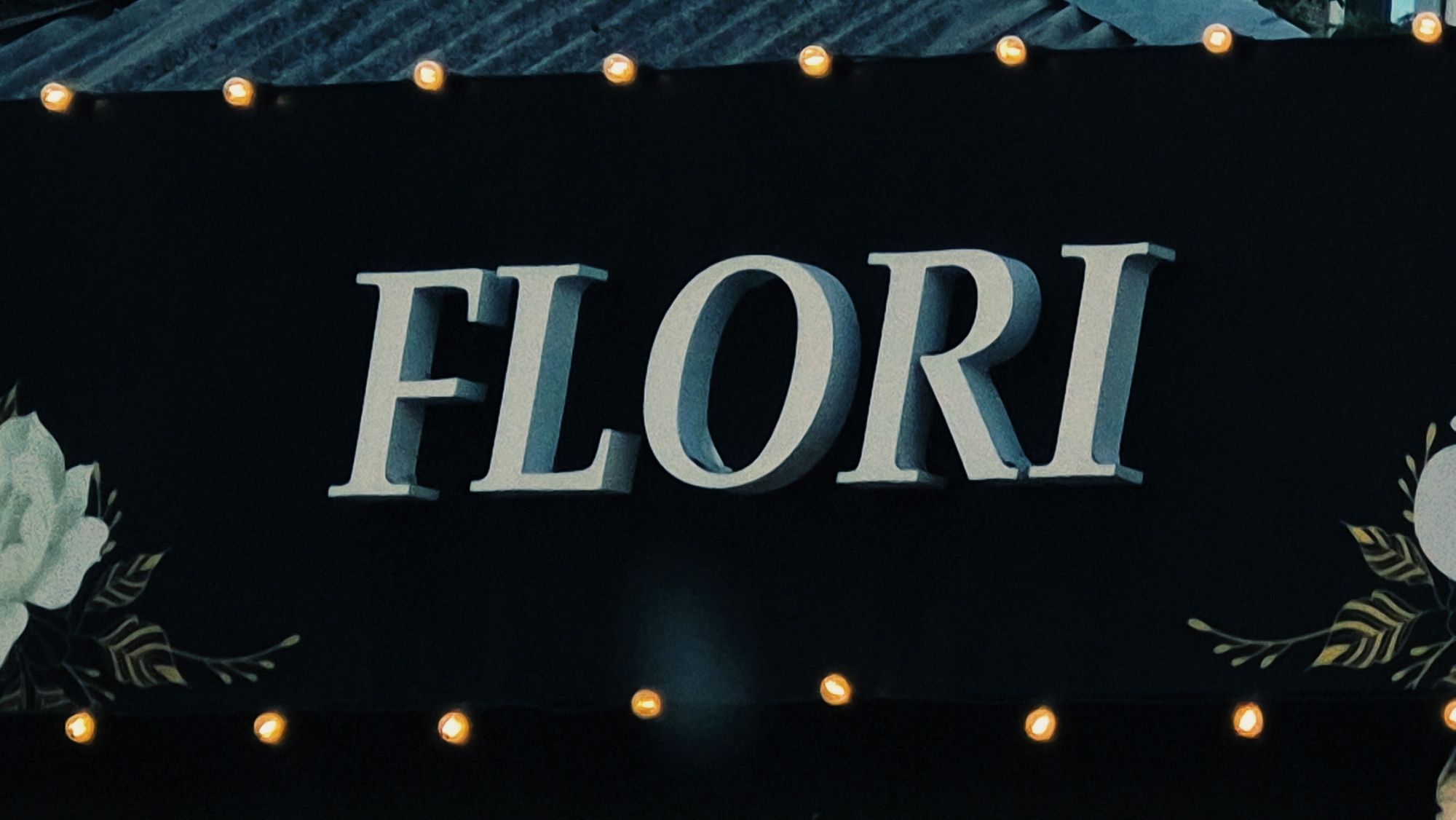 A sign with the word "FLORI" in large, 3D, light blue, serif letters is displayed against a dark background, possibly a black fabric or banner. The letters have a slightly weathered or textured appearance, with subtle shading that suggests they are three-dimensional. The sign is framed by a series of small, warm-toned lights, resembling fairy lights, strung along the top and bottom edges. To the left of the word is a floral design featuring a large, light-colored flower (likely a rose or peony) with detailed petals and dark green leaves and stems. To the right of the word, a similar floral motif is partially visible, with delicate leaves and stems. The overall aesthetic suggests a festive or decorative display, possibly for an event or establishment. The location in Chișinău, Moldova, is not explicitly visible in the image.