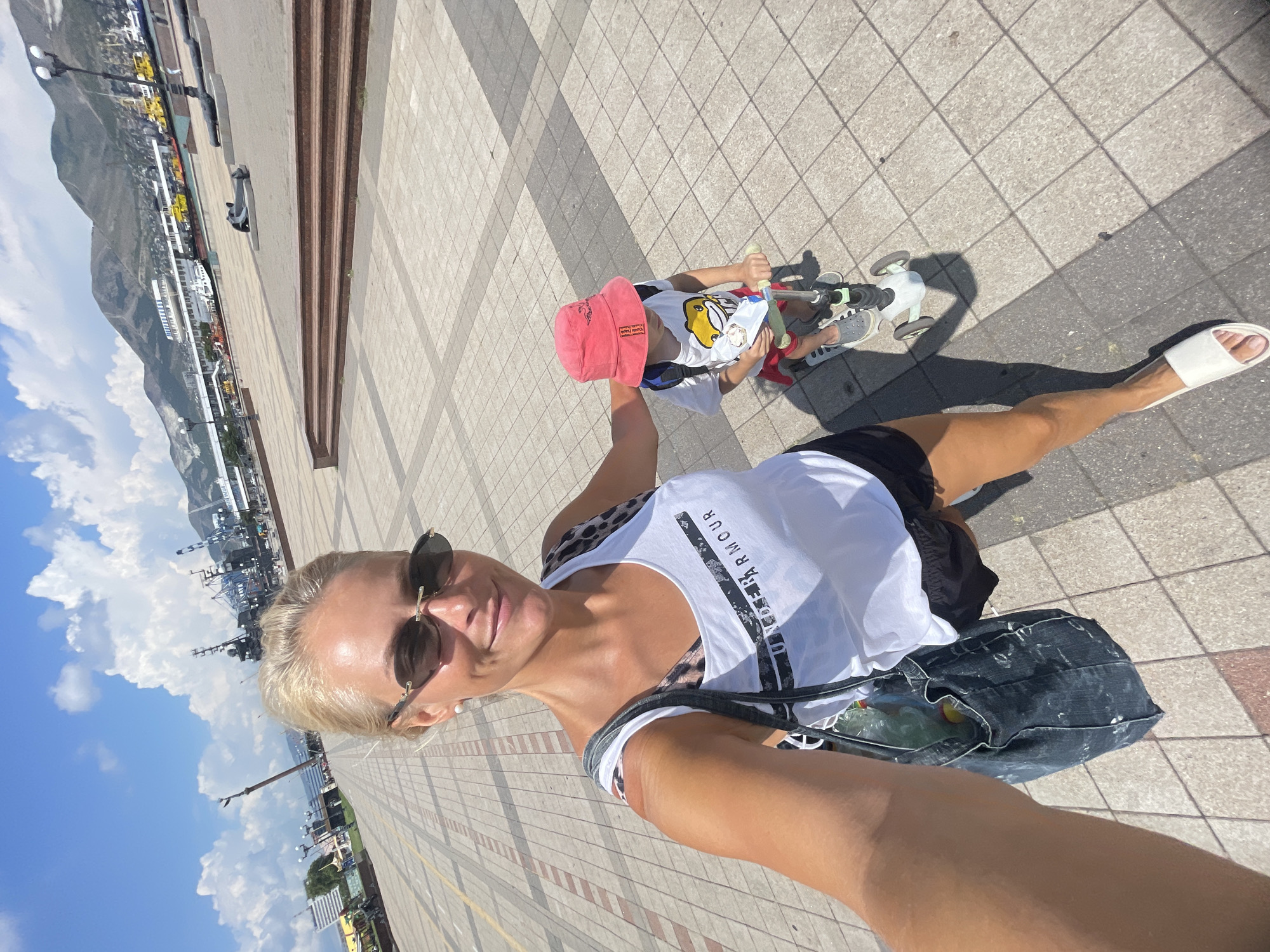 A woman and a child are on a paved promenade with a body of water and a city in the background under a sunny sky with clouds. The woman is in the foreground, taking a selfie. She has blonde hair, is wearing sunglasses, a white tank top with "UNDER ARMOUR" printed on it, and black shorts. She is carrying a large dark blue denim bag. To her right, a young child wearing a red bucket hat, a white t-shirt with a cartoon image, and red shorts is riding a scooter. The child is holding a white object. In the background, there is a harbor with ships, cranes, and buildings. Mountains are visible behind the city. The ground is paved with square tiles.