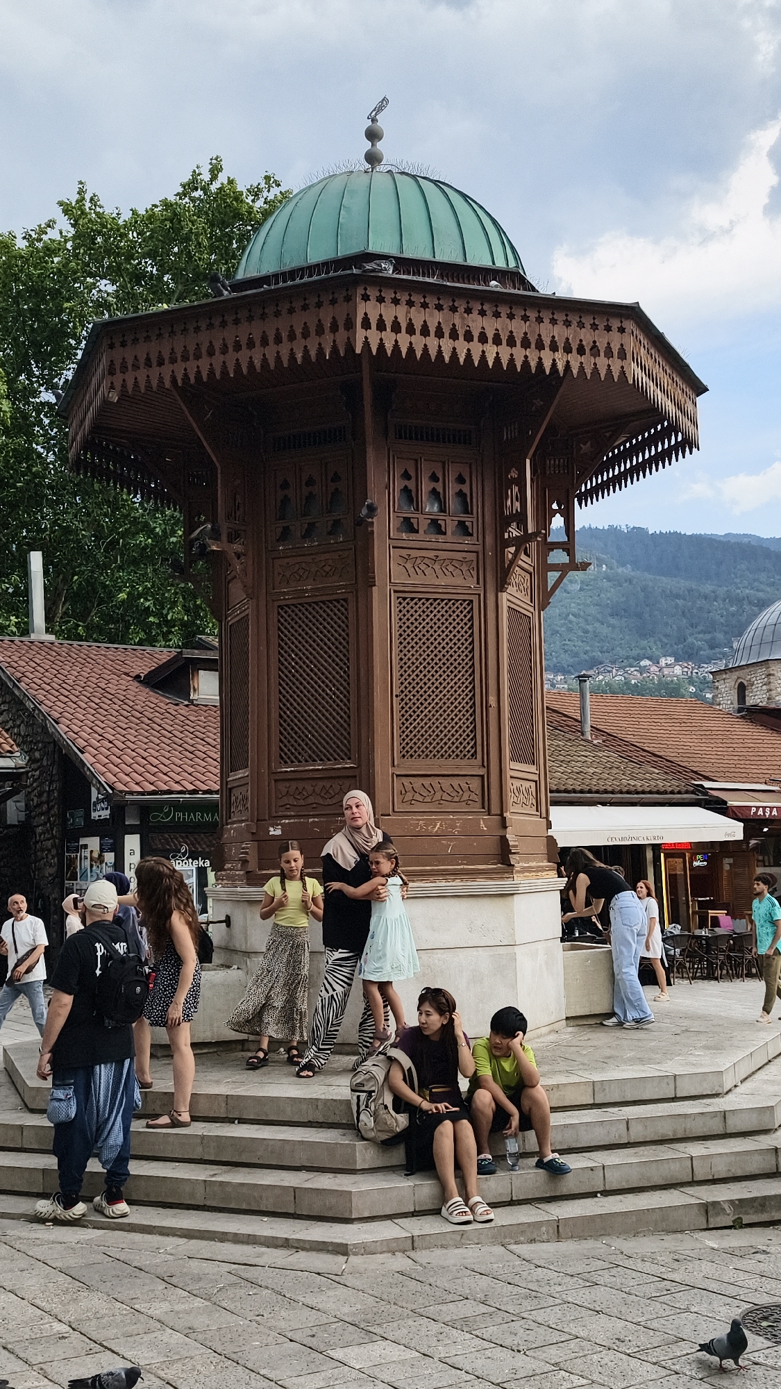 This is a street-level, medium-shot photograph of the Sebilj fountain in Baščaršija, Sarajevo, Bosnia and Herzegovina. The iconic wooden structure, featuring intricate carvings and a green copper dome, is the central focus. Several people are gathered around the fountain, some sitting on the tiered stone base, others standing.
On the left, a man wearing a black t-shirt and blue pants, carrying a backpack, stands facing the fountain. To his right, a woman with long brown hair is also standing.
In the middle of the frame, a woman in a beige hijab and black dress embraces two young girls. One girl, in a yellow shirt and patterned skirt, stands beside them, while the other, in a light blue dress, is held by the woman. To their right, a young woman in a black tank top and light blue jeans is bending down. Further to the right, a man in a turquoise shirt is walking away.
On the lower right side of the stone steps, a young woman with dark hair and sunglasses sits next to a boy in a lime green t-shirt. Both are looking in different directions.
The background reveals more of Sarajevo's characteristic architecture, with red-tiled roofs and the hilly landscape of the city. The sky is partly cloudy. Pigeons are visible on the fountain structure and on the cobblestone ground in the foreground. The scene captures a typical day at this popular tourist landmark.