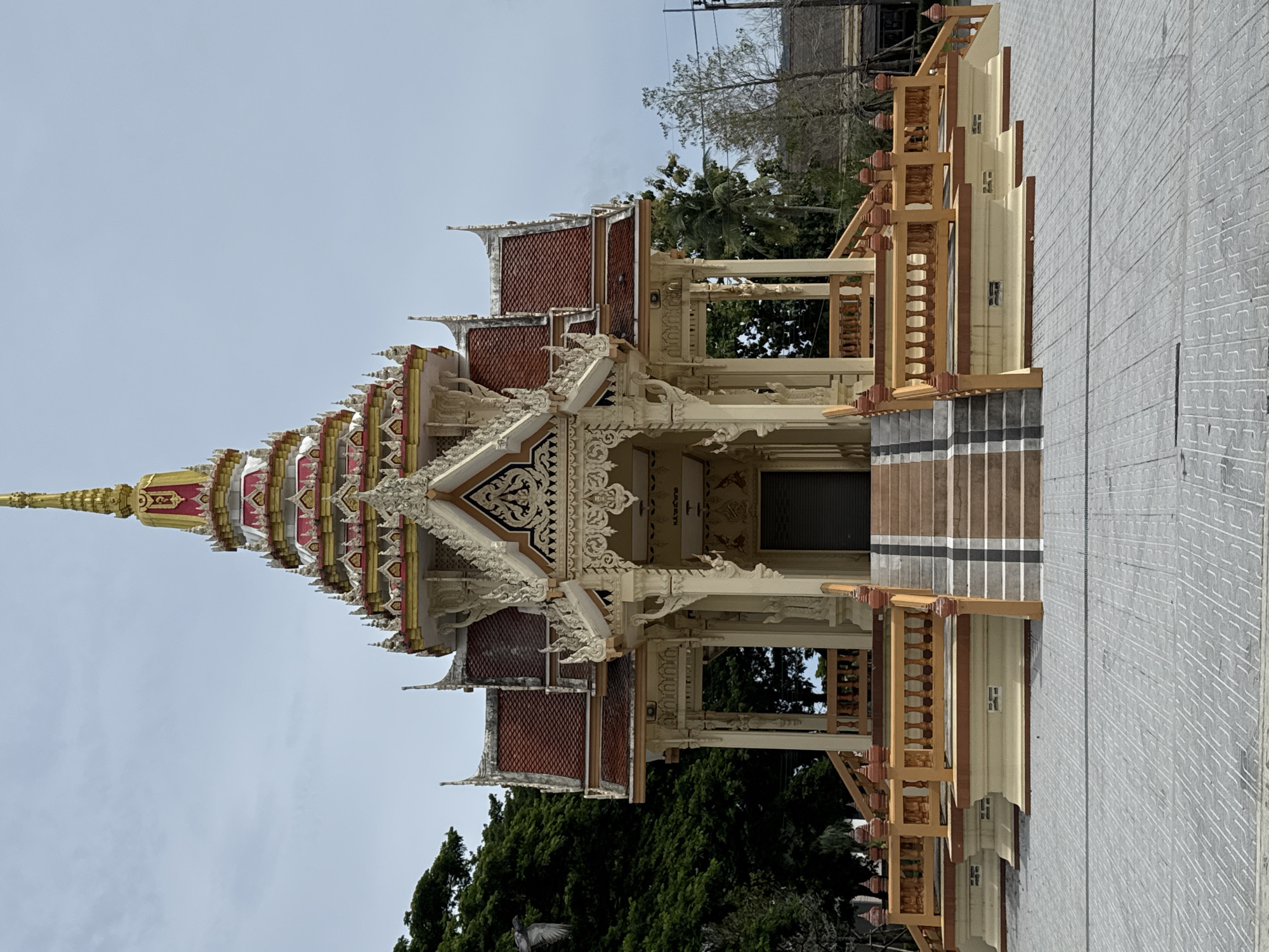 The image depicts the exterior of a crematorium in Chalong, Thailand. The structure is ornately decorated with a tiered, conical roof topped with a golden spire. The facade is painted in light yellow and cream with red accents on the roof tiles. A grand staircase with wooden railings leads up to the entrance, which is a dark doorway. The surrounding area is paved with grey tiles, and lush green trees are visible on the left side of the image. The sky is overcast.