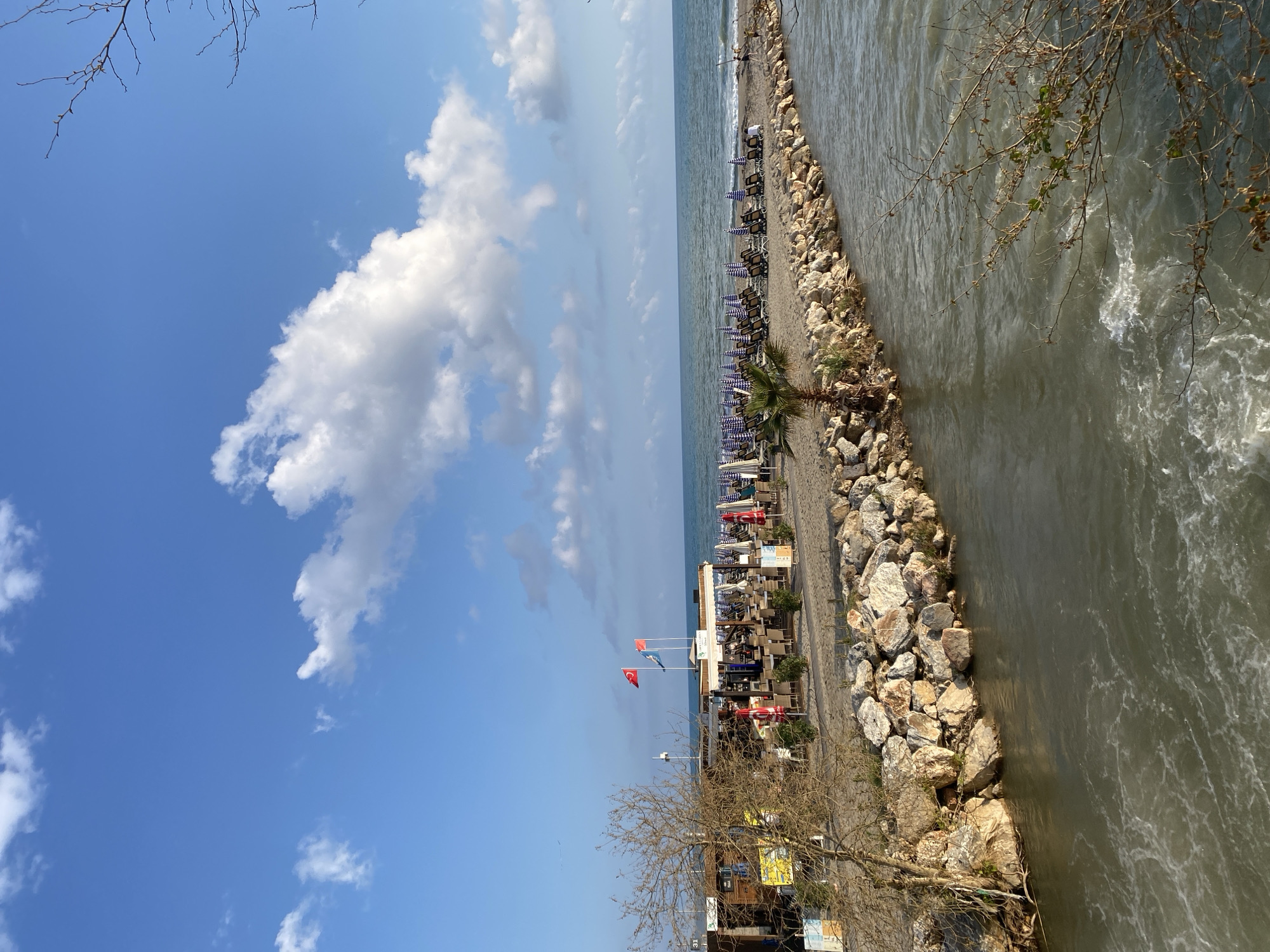 The image shows a beach scene in Alanya, Turkey. A waterway, possibly a river or canal, runs parallel to the beach, separated by a rock barrier. The beach is lined with lounge chairs and umbrellas. A small palm tree stands on the beach. A beachside establishment, marked by Turkish flags, is located closer to the foreground. The sky is blue with fluffy white clouds. Bare trees are visible in the foreground.