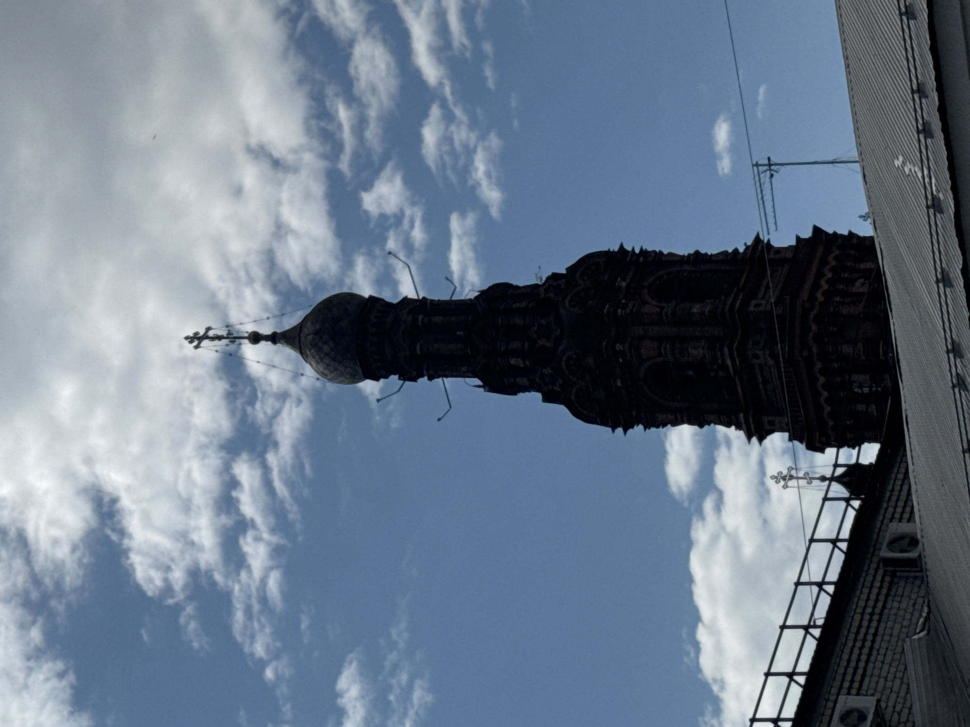 The image captures a tall, ornate church spire against a backdrop of blue sky and scattered clouds. The spire, likely part of an Orthodox church, is dark in color and displays intricate architectural details. It is topped with a dome and a cross. A building's edge, with exposed air conditioning units and metal railings, encroaches into the frame on the right. Given the architectural style, this is likely the Cathedral of St. Peter and Paul, located in the city of Kazan, Russia.
