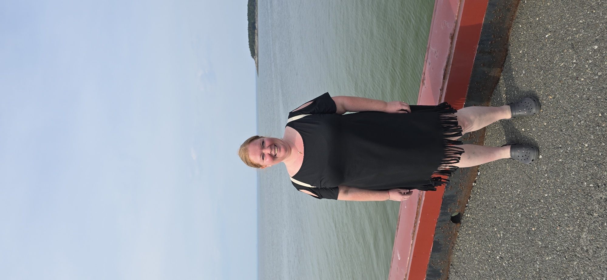 A medium shot captures a woman with blonde hair, wearing a black fringed dress and dark gray Crocs, standing on a gravel surface next to a red railing. The woman is smiling and facing the camera. Behind her, the ocean stretches out under a pale blue sky. In the distance, a small, tree-covered island is visible. The scene appears to be a waterfront location in Trois-Pistoles, Canada.