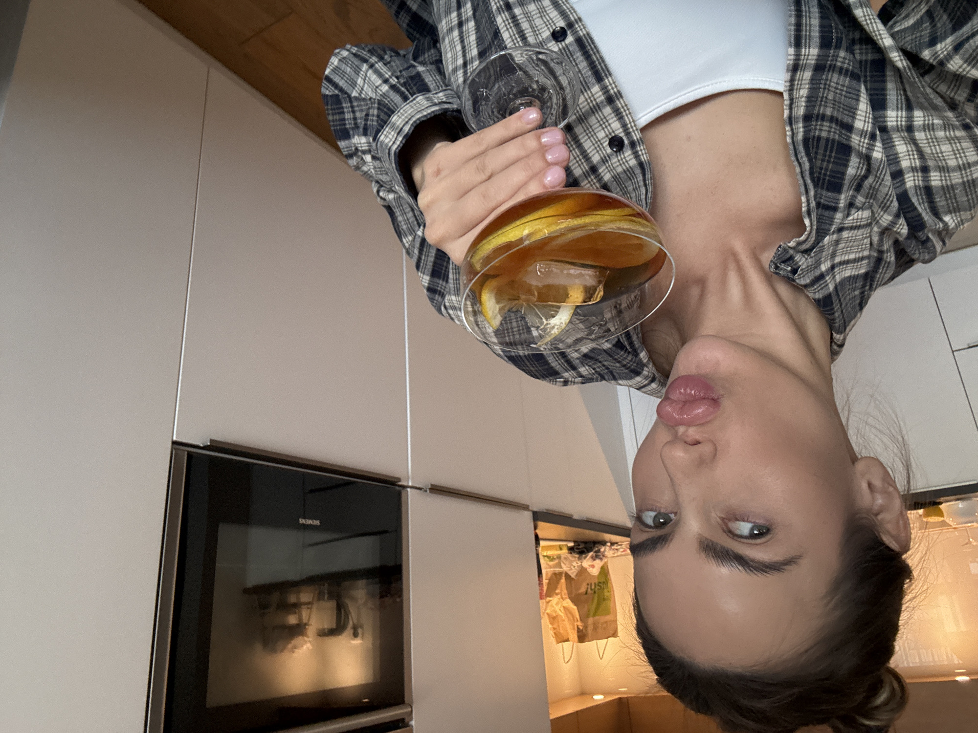 A woman is holding a cocktail glass in her hand. The glass contains ice and lemon. The woman is wearing a white shirt under a plaid flannel. The woman is in a kitchen with white cabinets and a Siemens oven. A shopping bag with the word "JUST" is visible in an open cabinet. The location of the image is in the city of Warsaw, Poland.