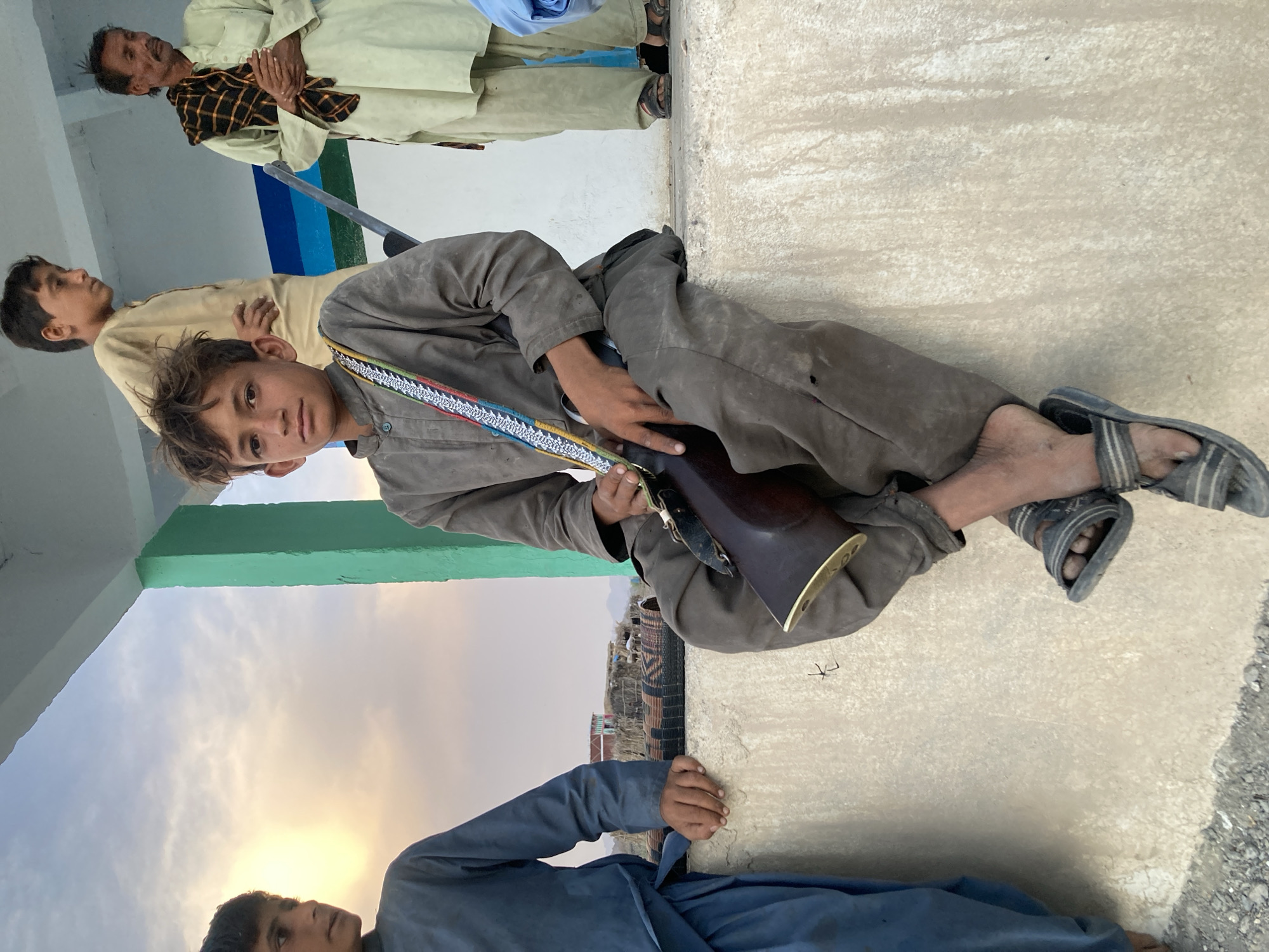 A medium shot depicts a group of people, including a young person holding a rifle, against a backdrop of what appears to be a cityscape or settlement, likely Wayara, Pakistan. The young person is dressed in a brown garment with a patterned strap, and is seated. The rifle is dark in color with metallic accents. Other individuals are visible in the background, some dressed in traditional attire. The location appears to be an outdoor, possibly elevated, area with visible building structures and sky.