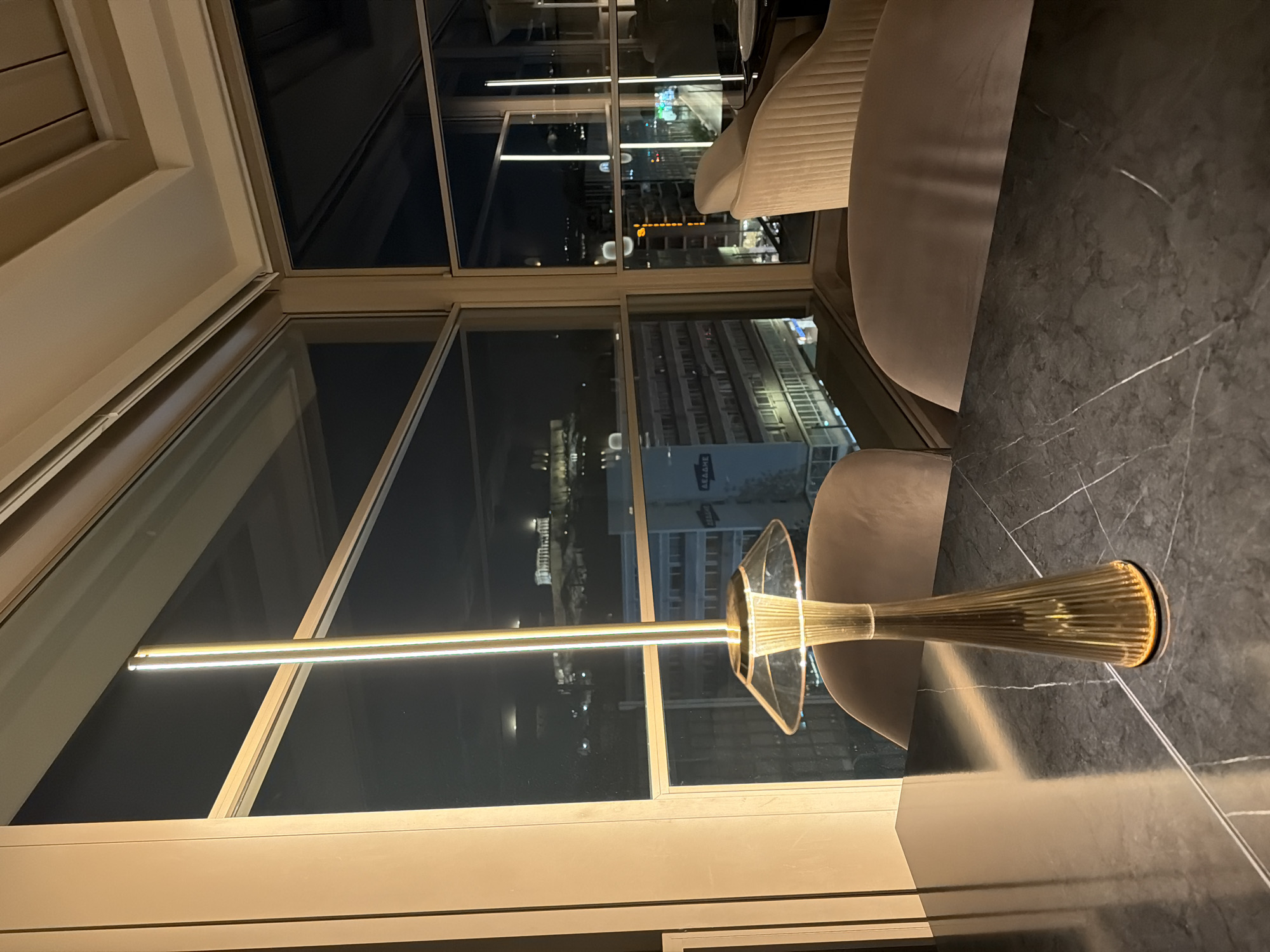 This image, originally rotated 90 degrees clockwise, captures a modern interior looking out over a cityscape at night. In the foreground, a dark, veined marble surface, likely a table or counter, occupies the right side. On the left, a prominent, contemporary floor lamp features a golden, fluted base tapering upwards, topped with a clear, geometric shade. It emits a bright, vertical line of light, reflecting subtly on the surrounding surfaces.
Several light-colored, possibly velvet-upholstered, modern chairs are partially visible in the mid-ground, suggesting a chic dining or lounge area. The interior is framed by large windows with light-colored sills and frames, offering a panoramic view of the city.
Beyond the windows, the cityscape is illuminated by numerous artificial lights against the dark night sky. Buildings with glowing windows and streetlights create a vibrant urban tapestry. On one building, partially visible text in Greek, which appears to be "ΑΒΒΑΔΕ", can be seen. In the distance, a significant ancient structure with visible columns, strongly resembling the Acropolis, is faintly lit, confirming the location as Athens, Greece. The overall ambiance is sophisticated and urban, characteristic of an upscale establishment offering night views.